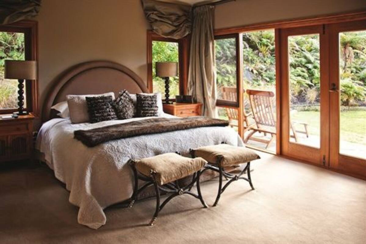 A bedroom with a large bed, two bedside tables, a bench, and a view of the outdoors through a sliding glass door.