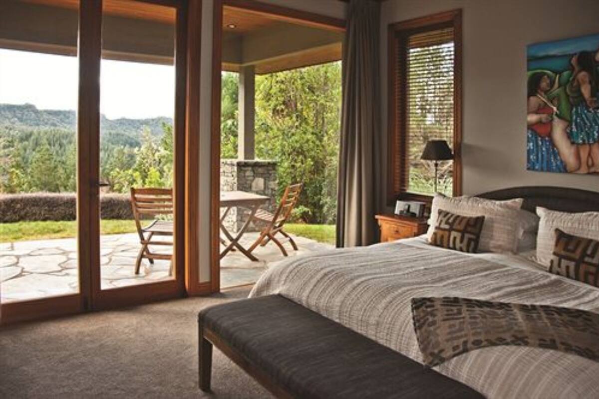 A bedroom with a large bed, a bench, a table, and chairs near a sliding glass door leading to a patio with a stone table and chairs.