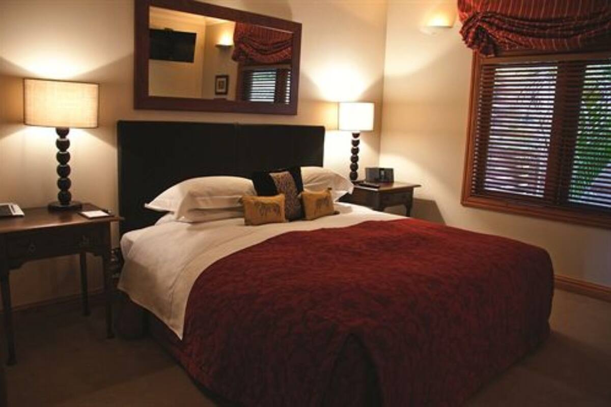 A neatly made bed with a red blanket, two bedside tables with lamps, a mirror above the bed, and a window with blinds.