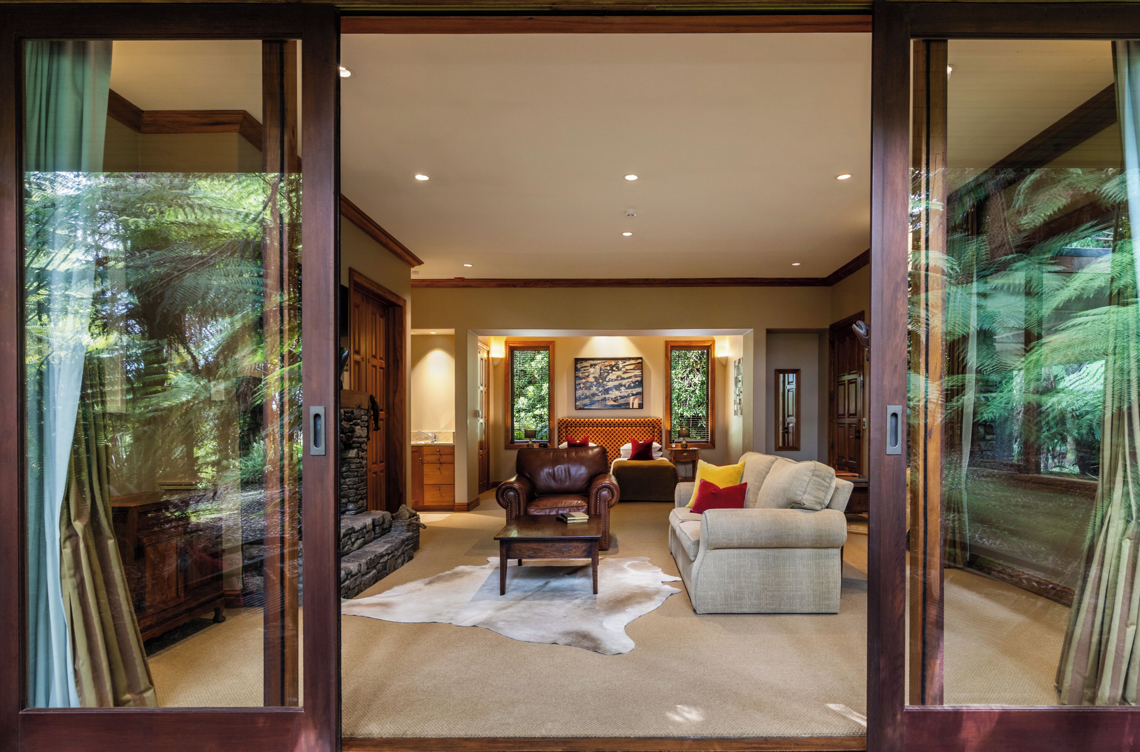 A spacious living room with a leather sofa, a grey couch, and a coffee table. There is a stone fireplace and a large window with a view of trees.