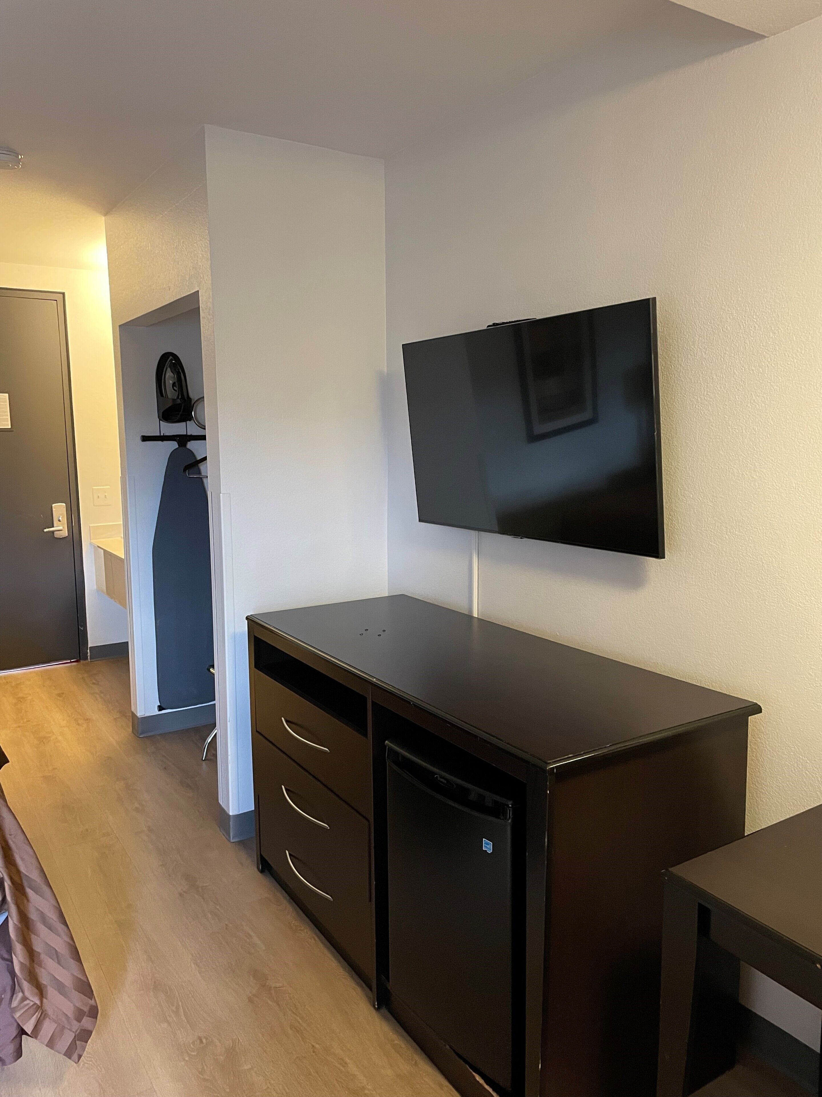 A room with a flat-screen TV mounted on the wall, a dark wooden desk with drawers, and a compact refrigerator.