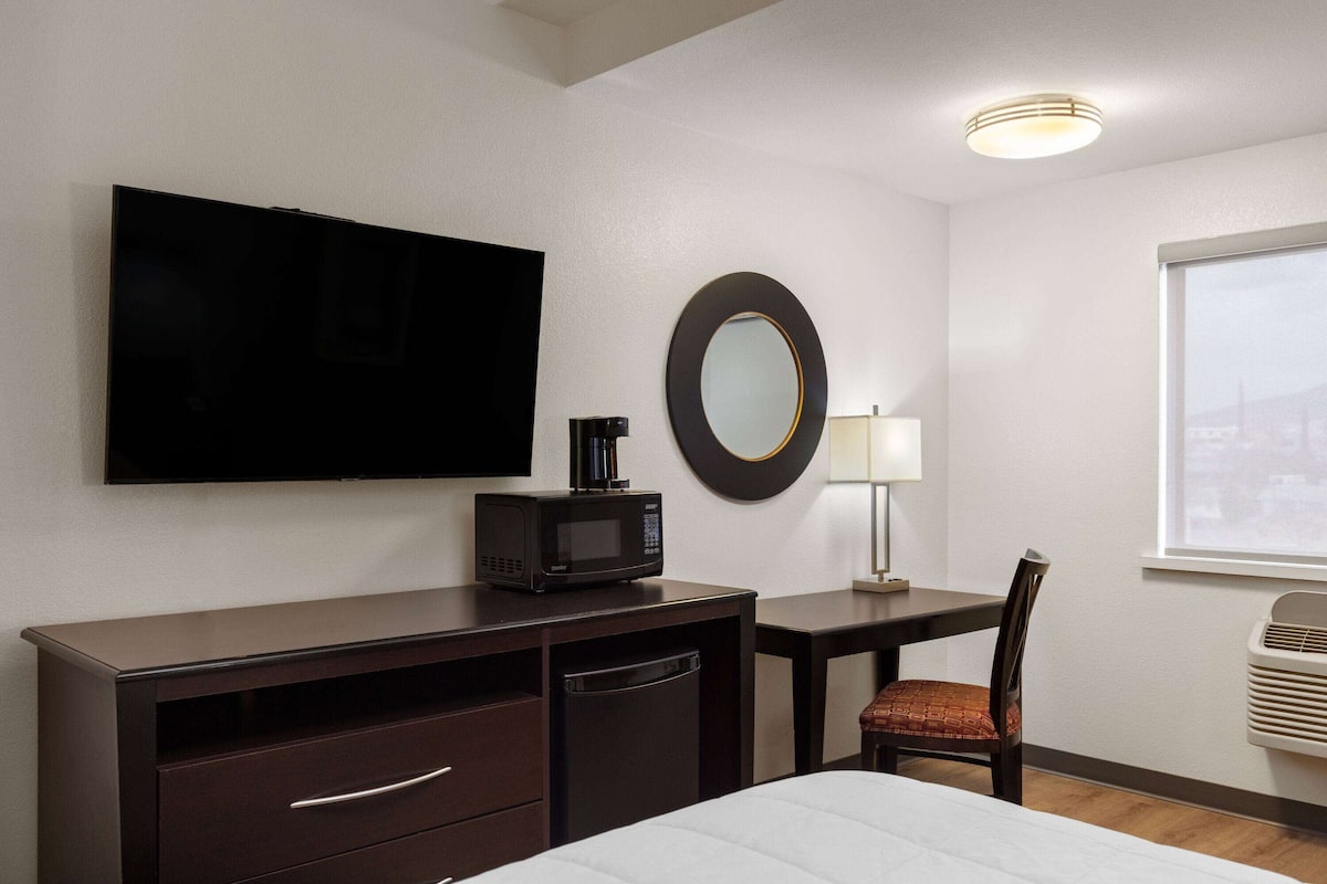 Ein Hotelzimmer mit einem Bett, einem Schreibtisch mit Mikrowelle, einem Stuhl, einem Fernseher, einem Spiegel, einer Lampe und einem Fenster.