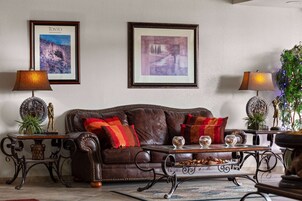 Uma sala de estar com um sofá de couro marrom, duas mesas laterais com luminárias, quadros nas paredes e uma planta decorativa.