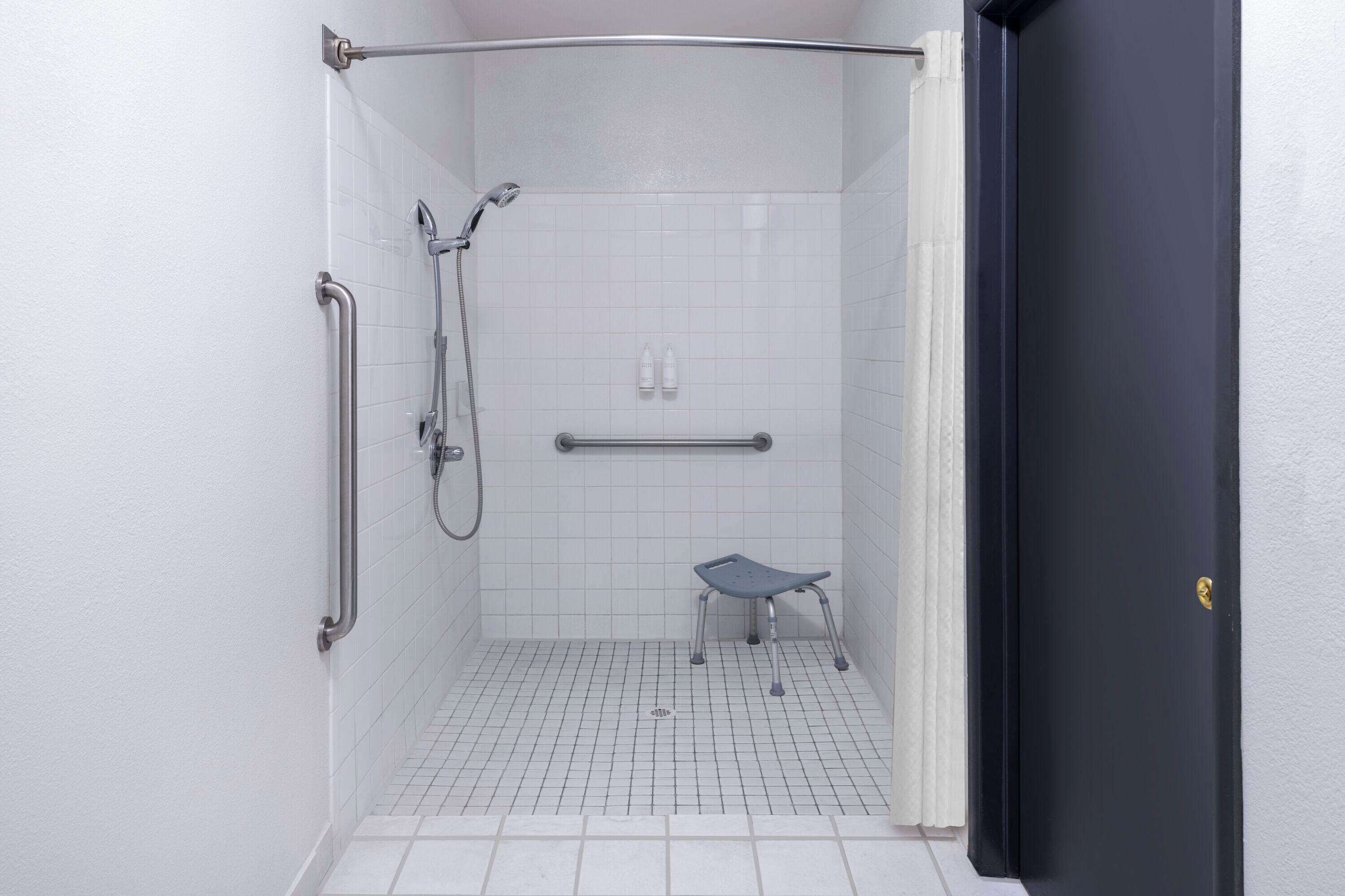Une petite salle de douche aux murs recouverts de carreaux blancs, équipée d’une barre d’appui, d’un pommeau de douche et d’une barre de maintien.