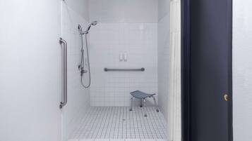 Une petite salle de douche aux murs recouverts de carreaux blancs, équipée d’une barre d’appui, d’un pommeau de douche et d’une barre de maintien.