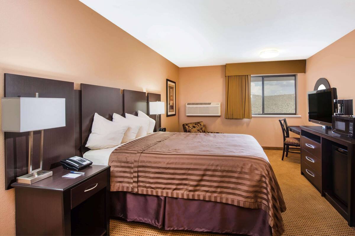 Ein Hotelzimmer mit einem Bett, einem Schreibtisch mit Fernseher, einem Sessel, einer Schreibtischlampe, einem Telefon und einer Klimaanlage.