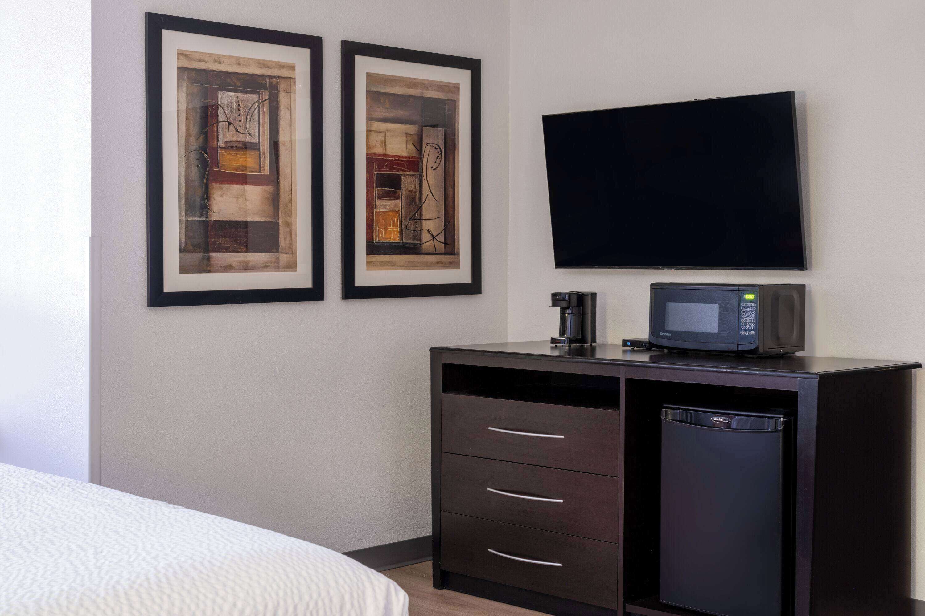 A hotel room with a bed, a dark wooden dresser with a microwave and a small refrigerator, and two framed abstract paintings on the wall.