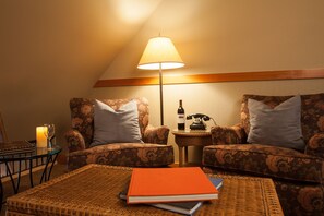 A cozy room with two floral-patterned armchairs, a small side table with a bottle and a telephone, and a wicker ottoman with books.