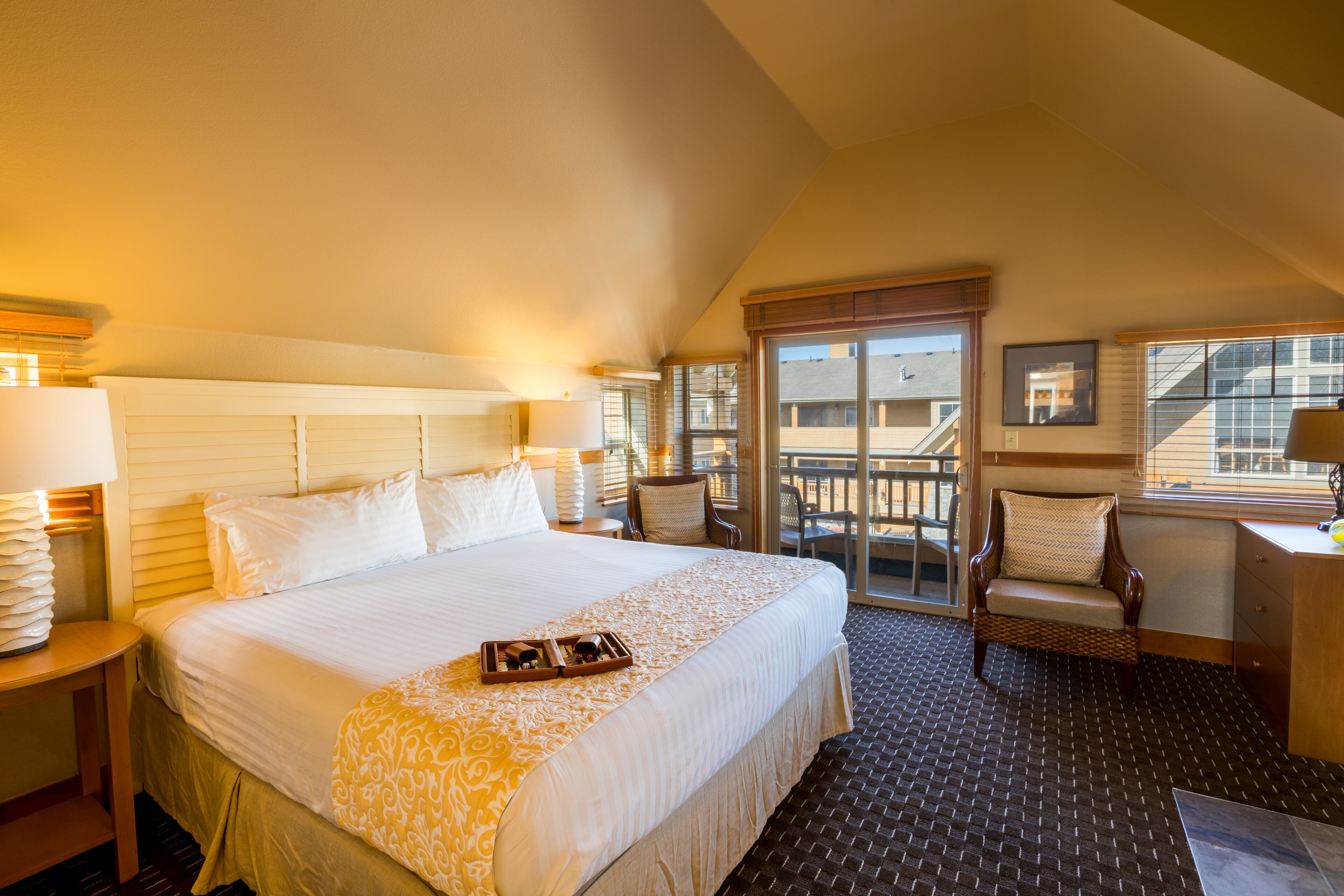 A hotel room with a large bed, a desk with a lamp, a chair, and a balcony with a view of the building.