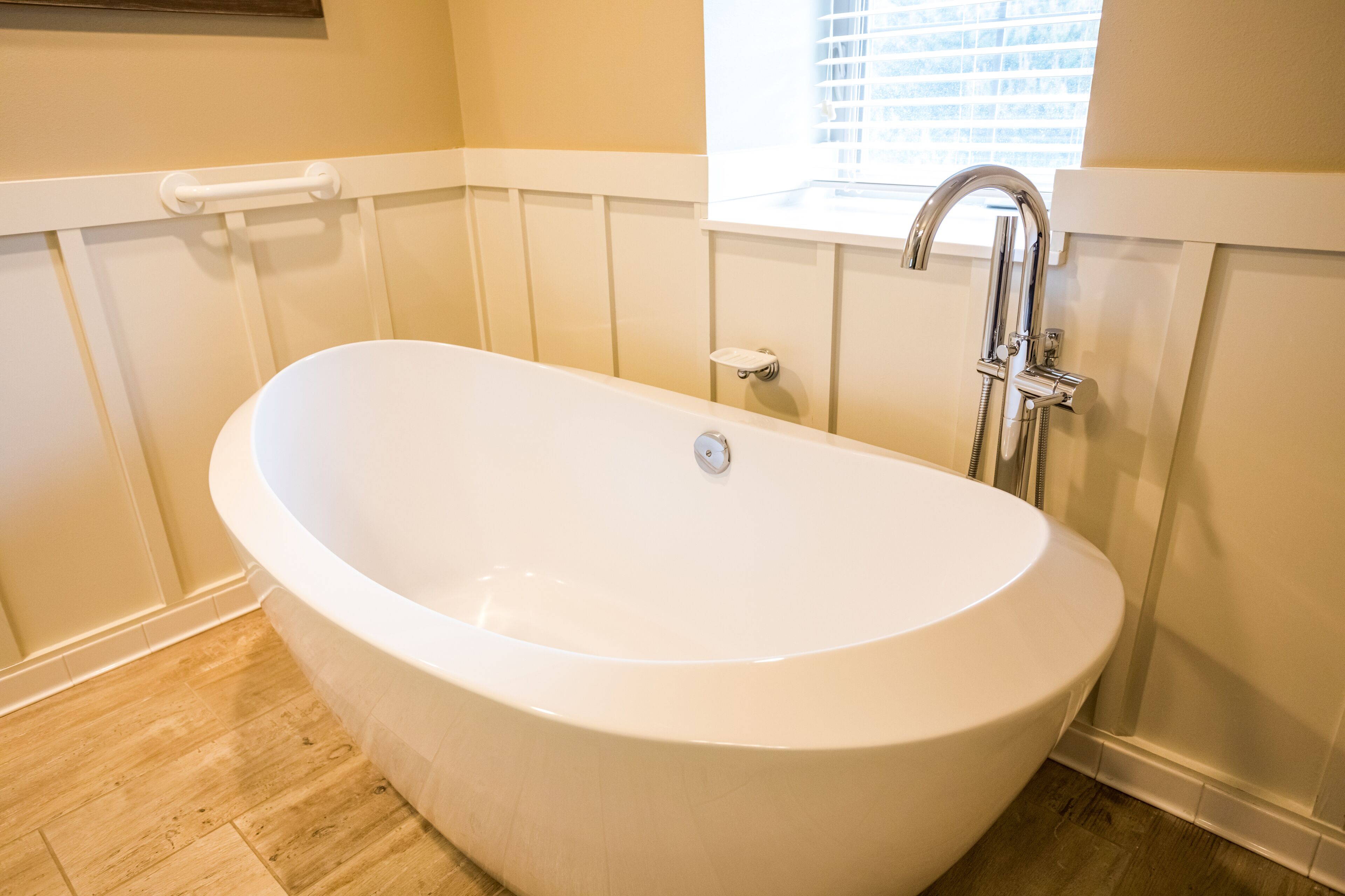 A modern bathroom with a freestanding bathtub, a window with blinds, and wooden flooring.