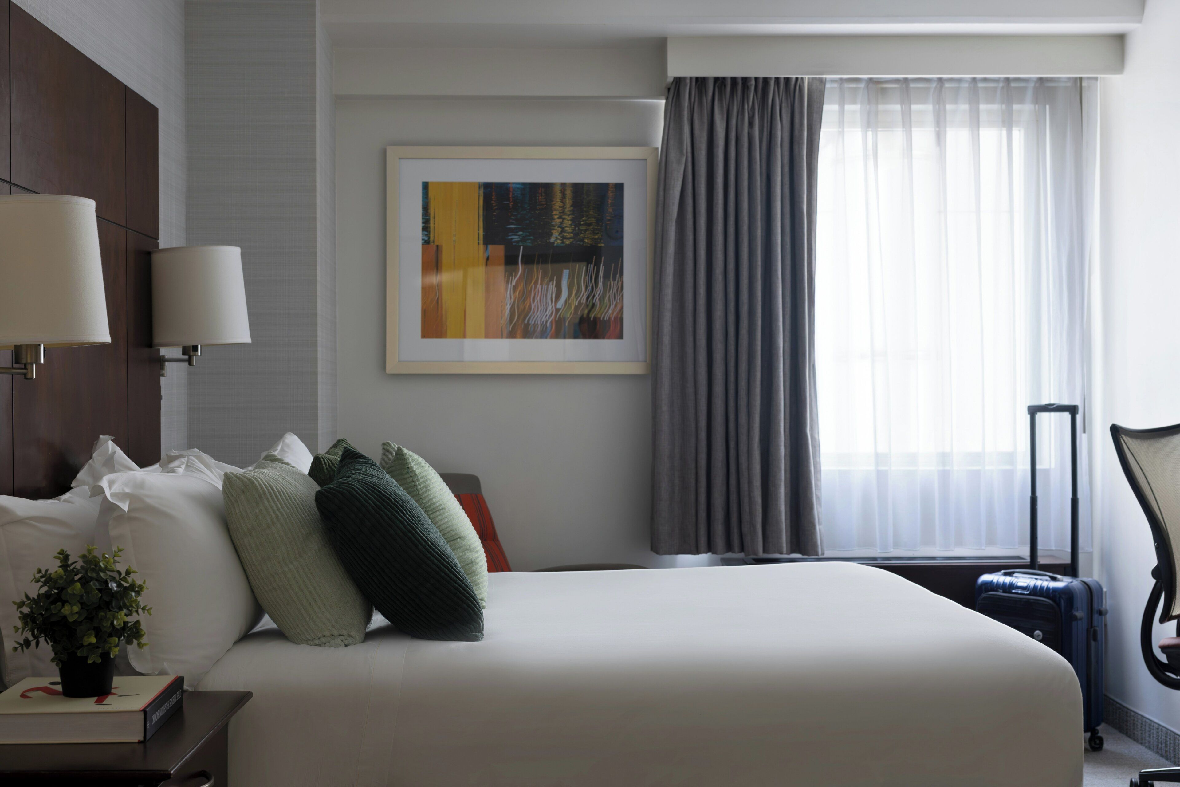 Quarto de hotel com cama, almofadas, abajur, uma obra de arte emoldurada, janela com cortinas e uma mala.