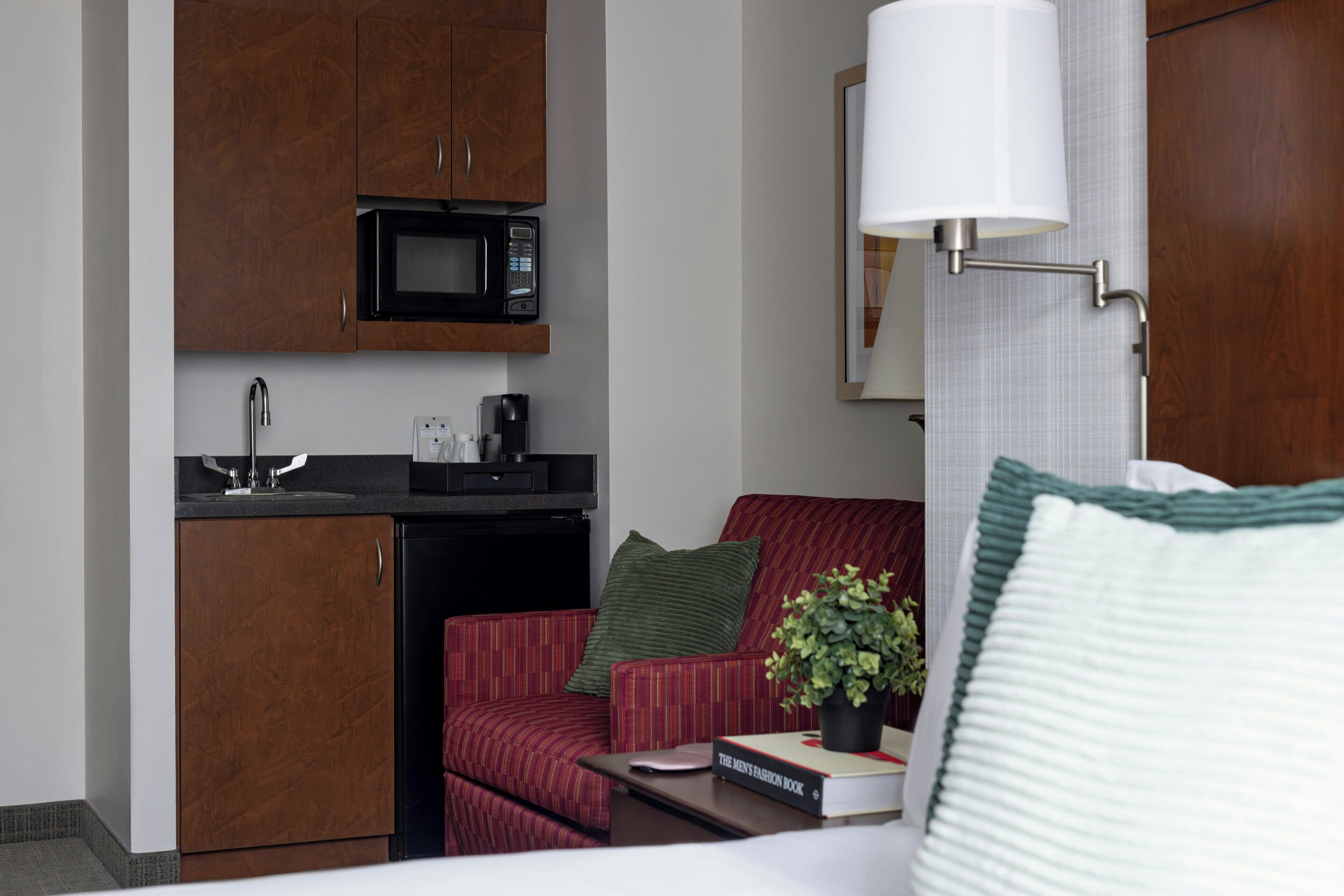 A compact hotel room with a kitchenette, a bed, a red armchair, a bedside table with a book and a plant, and a wall-mounted lamp.