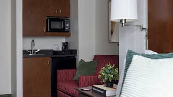 A compact hotel room with a kitchenette, a bed, a red armchair, a bedside table with a book and a plant, and a wall-mounted lamp.