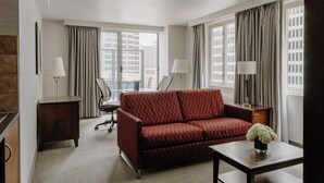 A hotel room with a red sofa, a desk with a lamp, a chair, and a view of buildings through large windows. - Club Quarters Hotel Embarcadero, San Francisco (San Francisco)