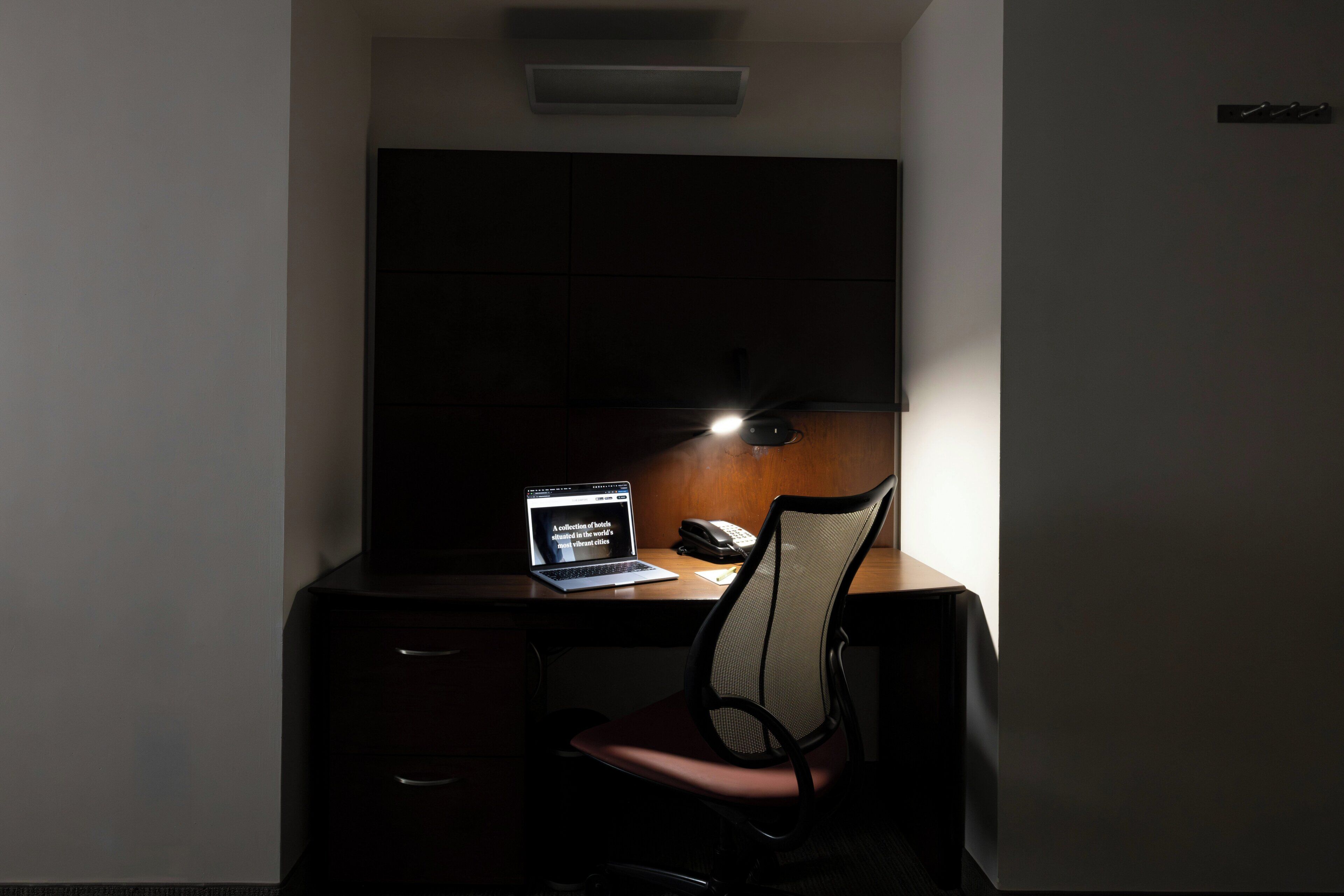Una habitación oscura con un escritorio, una silla y una computadora portátil mostrando un sitio web de hotel.