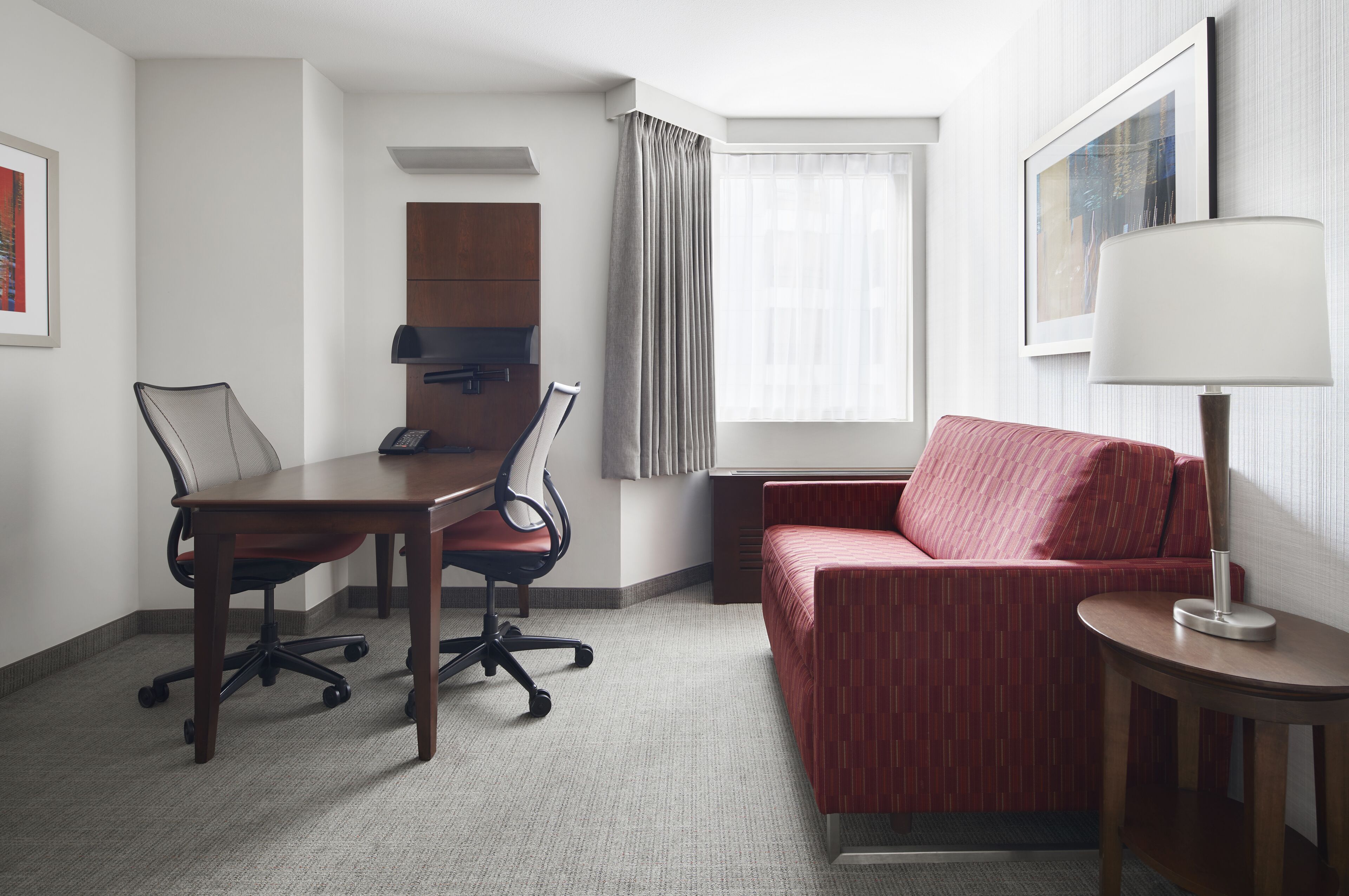 Et hotelværelse med et skrivebord, to stole, en rød sofa, en lampe og et vindue med gardiner.