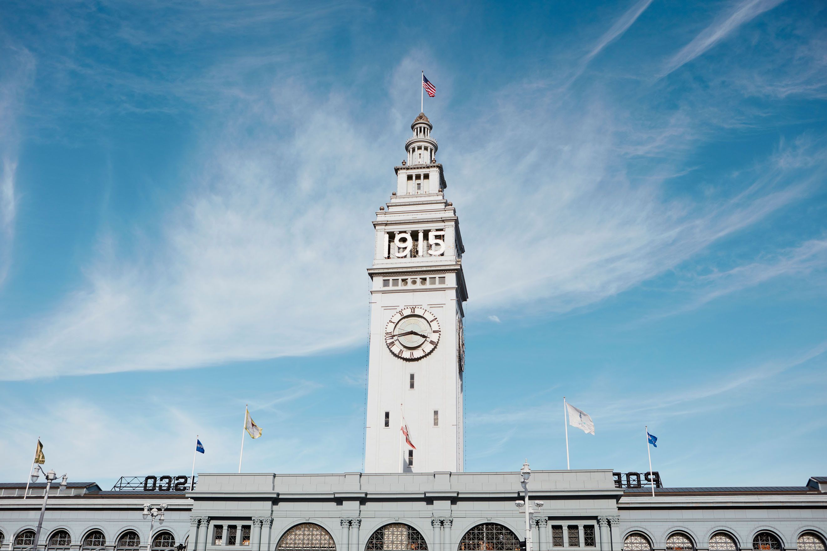 A tall clock tower with the year 1915, surrounded by a building with arched windows and flags.
