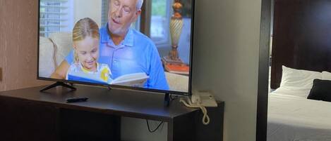 A television on a dark wooden desk in a hotel room, with a bed and a lamp in the background.