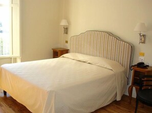 A neatly made bed in a room with a wooden nightstand, a lamp, and a chair. - Grand Hotel Certosa (Padula)