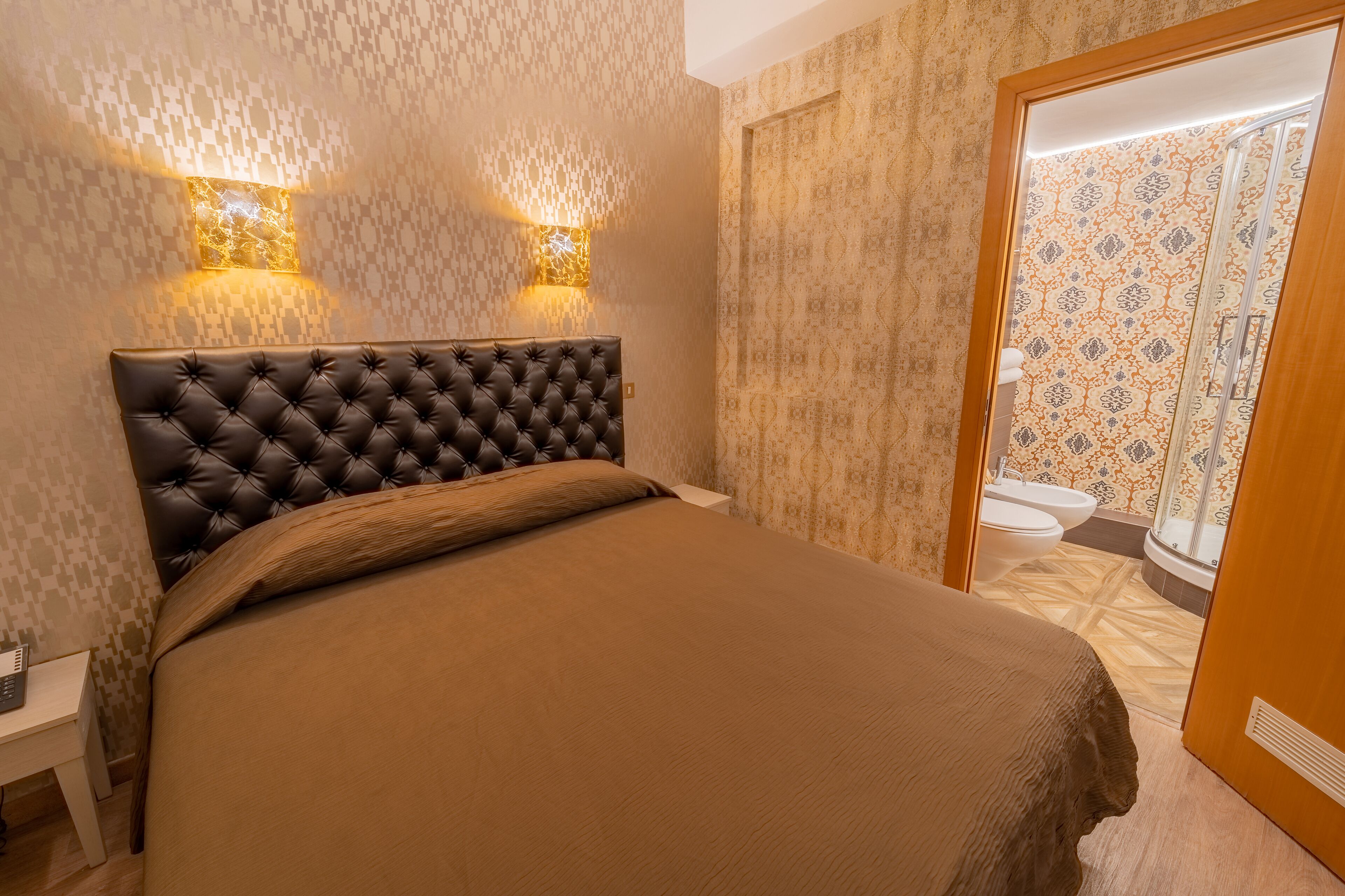 Een slaapkamer met een gestuikt hoofdeinde, een bed met een bruine deken, twee wandlampen en een badkamer die door een open deur zichtbaar is.