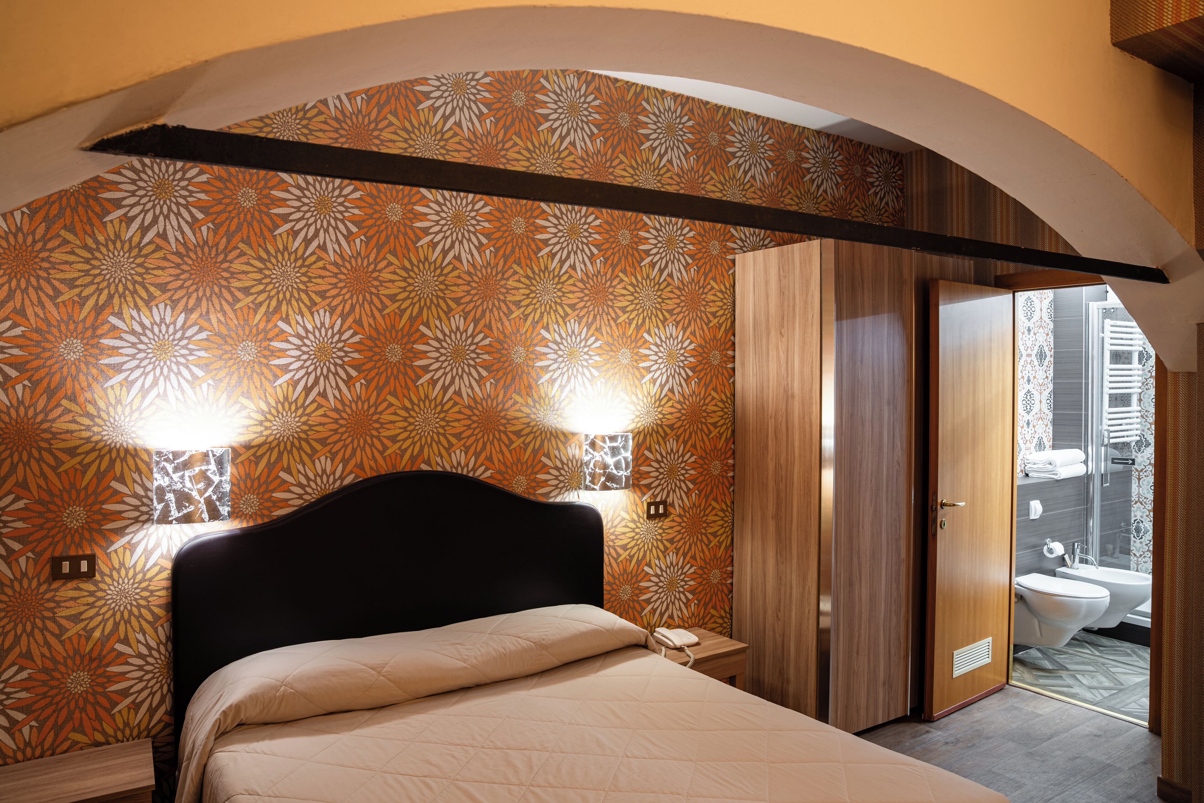 A bedroom with a bed, a nightstand, a wall-mounted lamp, and a door leading to a bathroom.