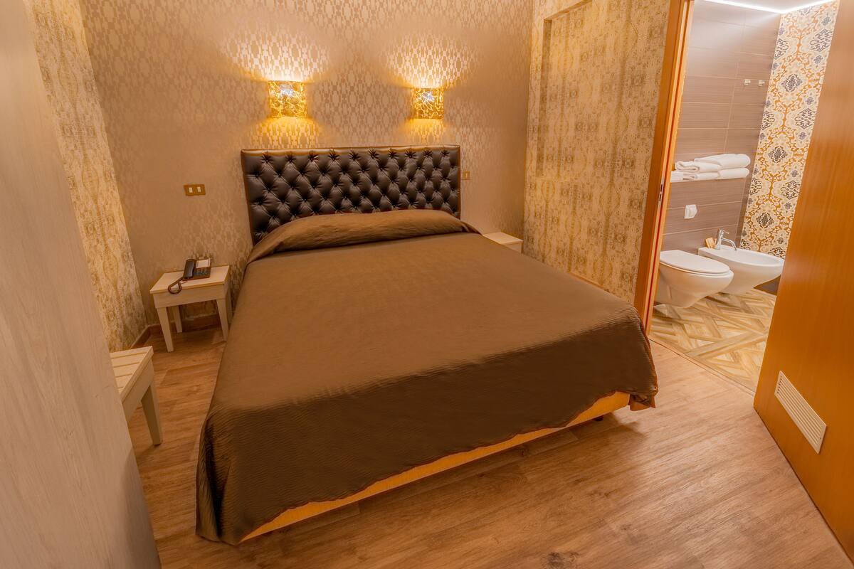 A bedroom with a large bed, a bedside table with a lamp, and a bathroom visible through an open door.