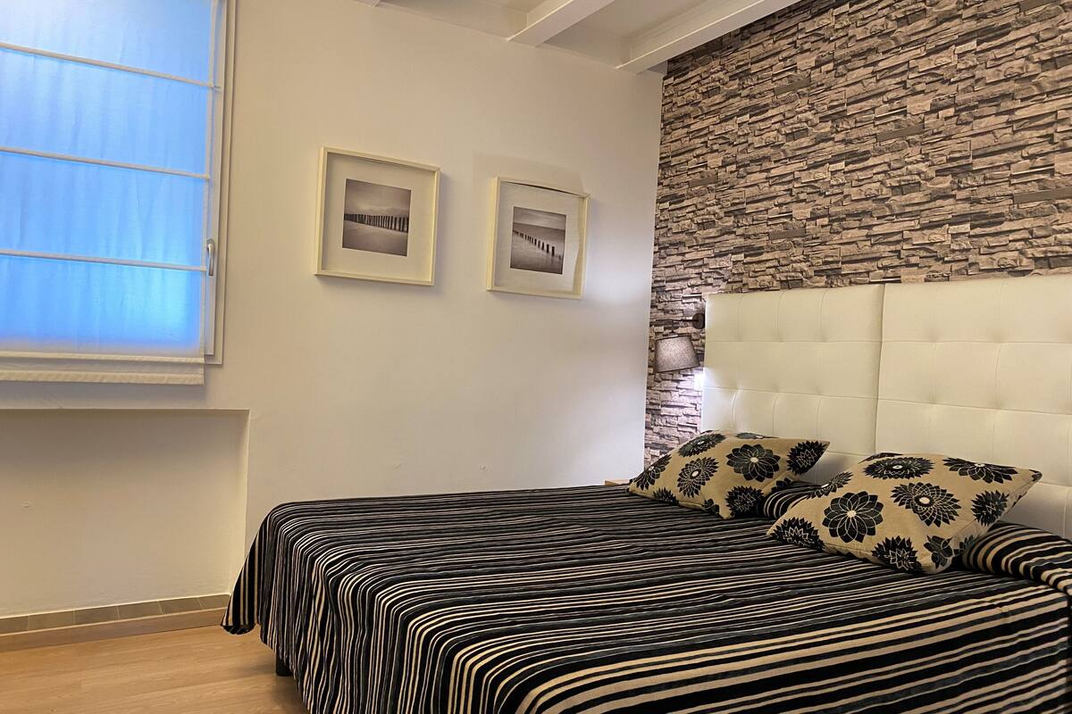 A bedroom with a bed, two framed pictures on the wall, a window with blinds, and a stone-textured wall.