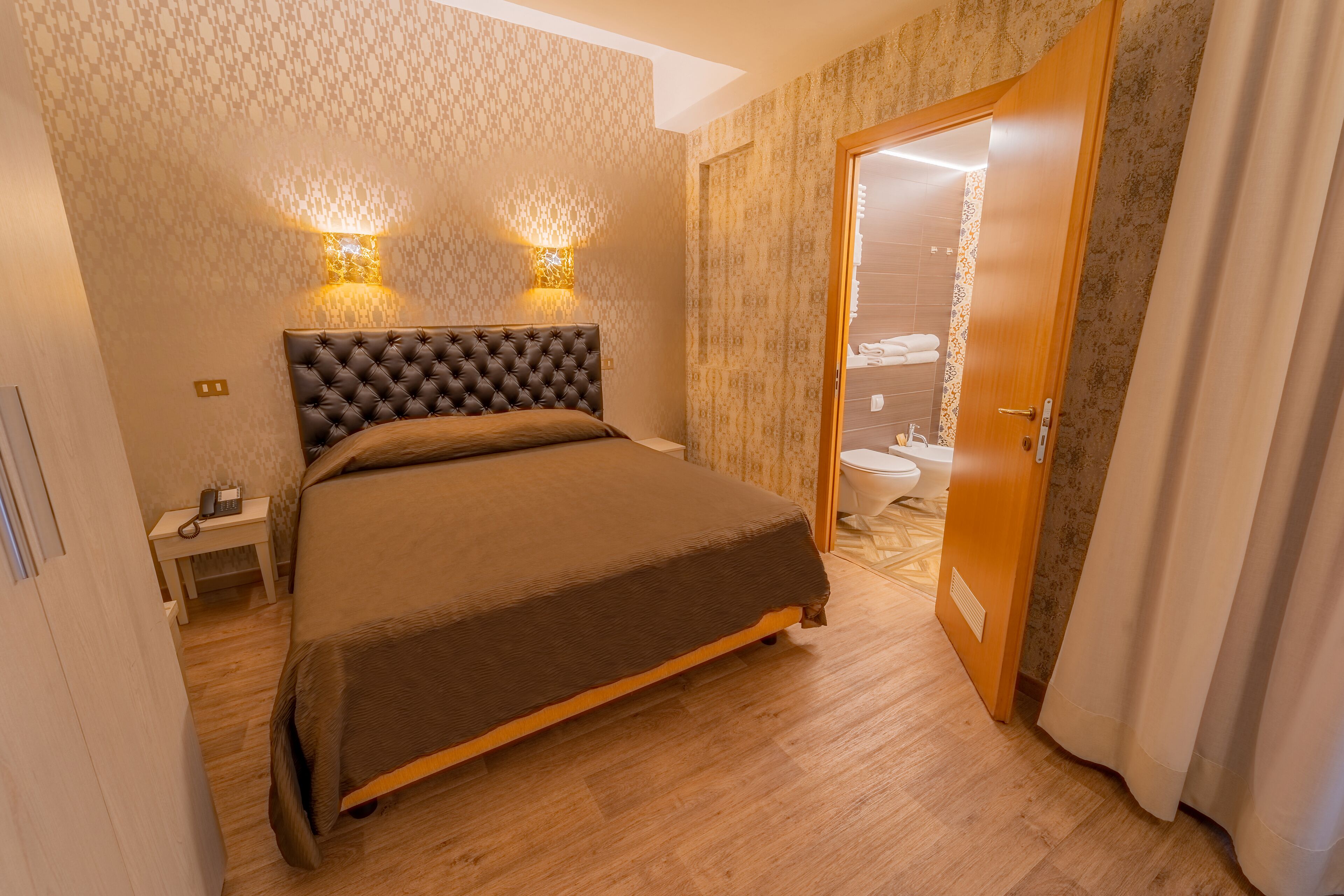 Une chambre d’hôtel avec un grand lit, deux lampes fixées au mur, une table de chevet avec un téléphone et une porte en bois donnant sur une salle de bain.