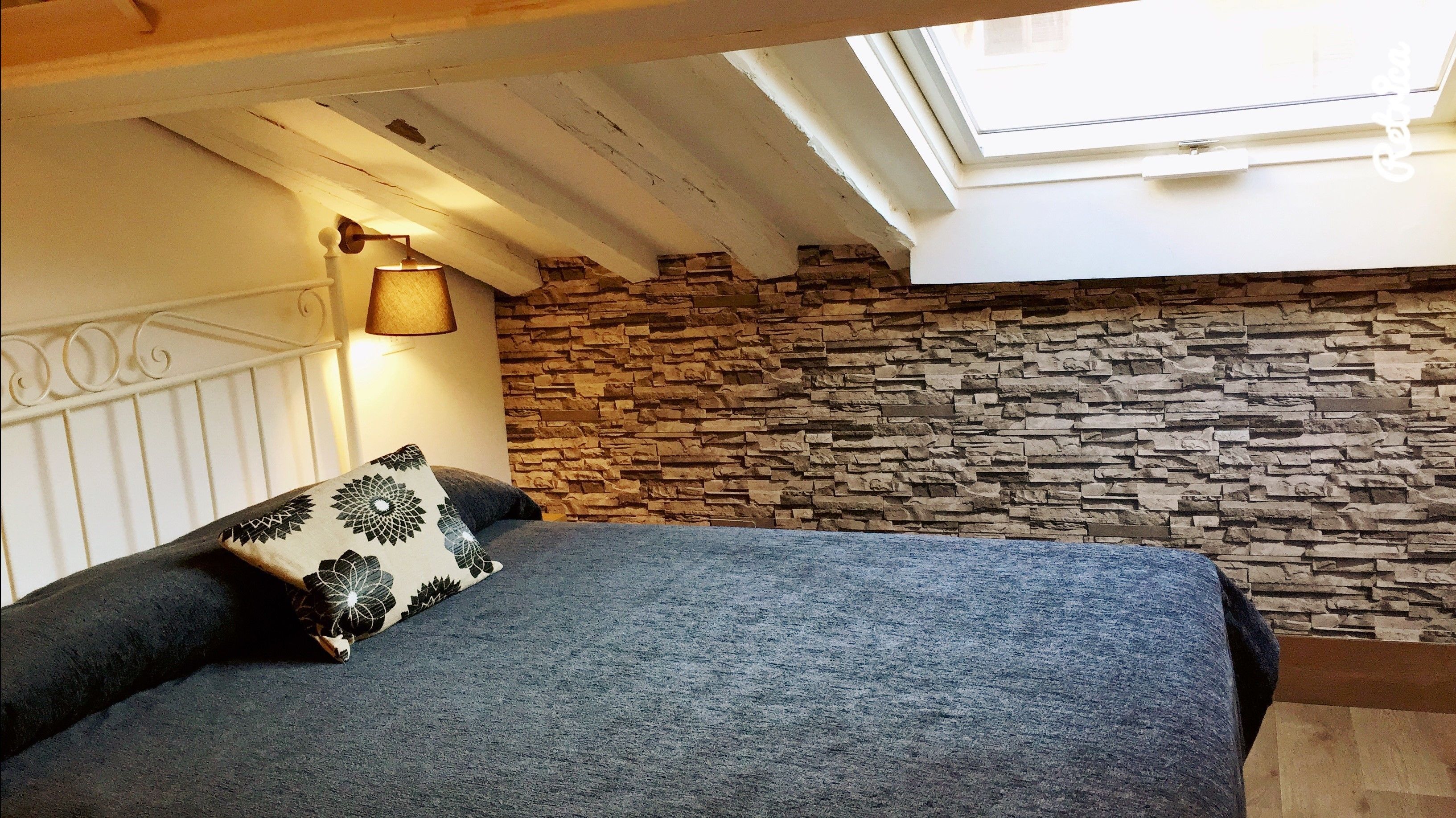 Ein Schlafzimmer mit Steinwand, Dachfenster und einem Bett mit gemusterten Kissen.