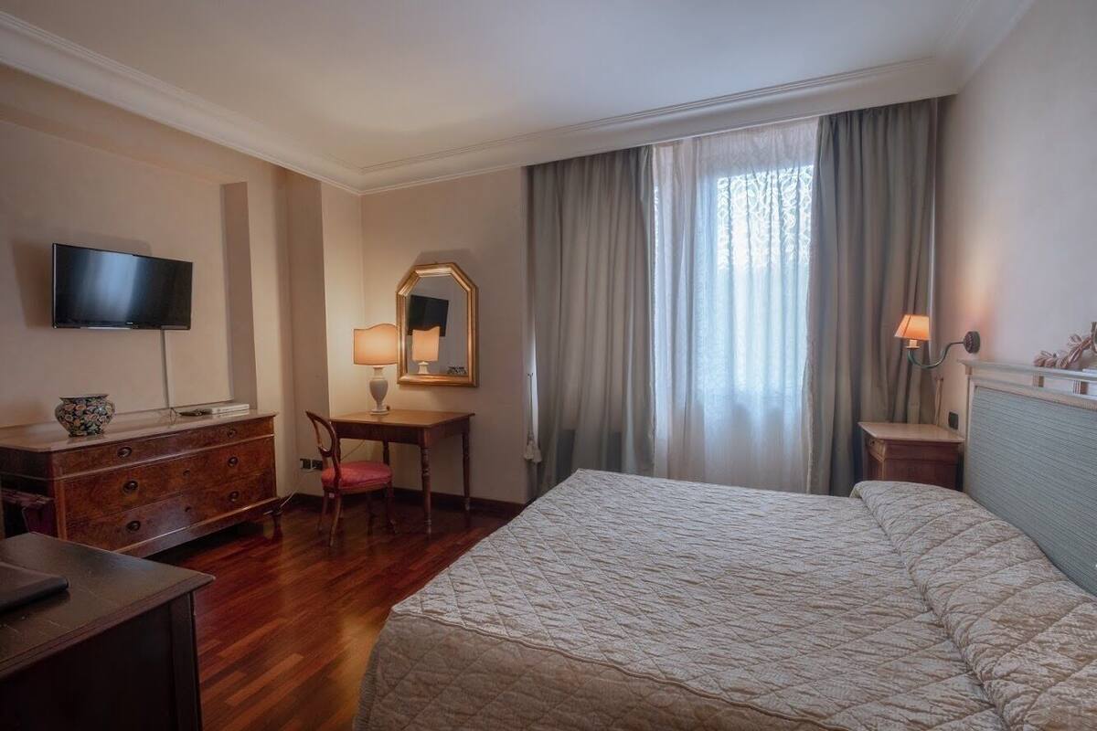 A hotel room with a bed, a desk, a chair, a television, a dresser, and a window with curtains.