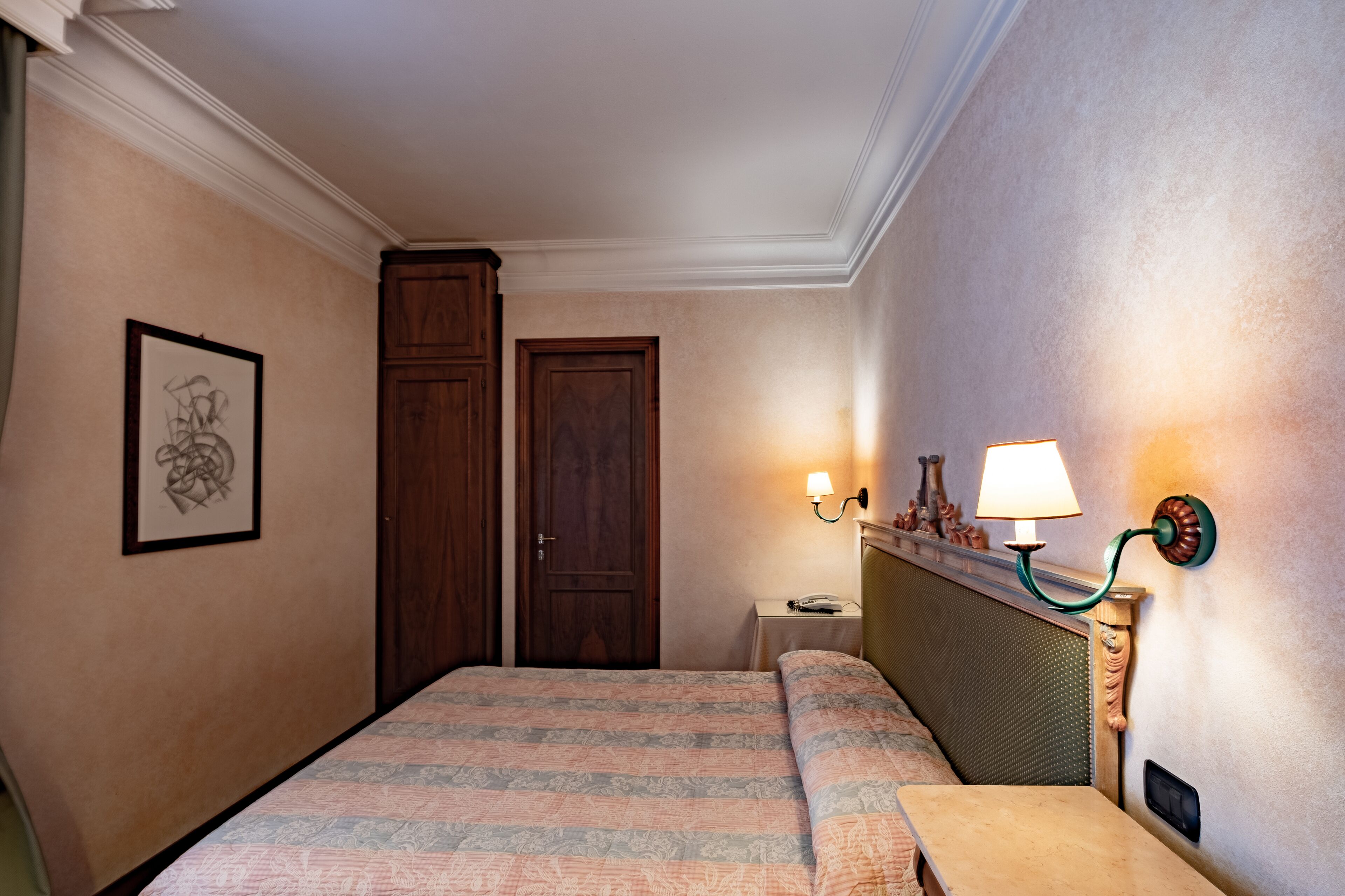 Una camera d'albergo con un letto, un comodino, una lampada, un quadro appeso alla parete e una porta.
