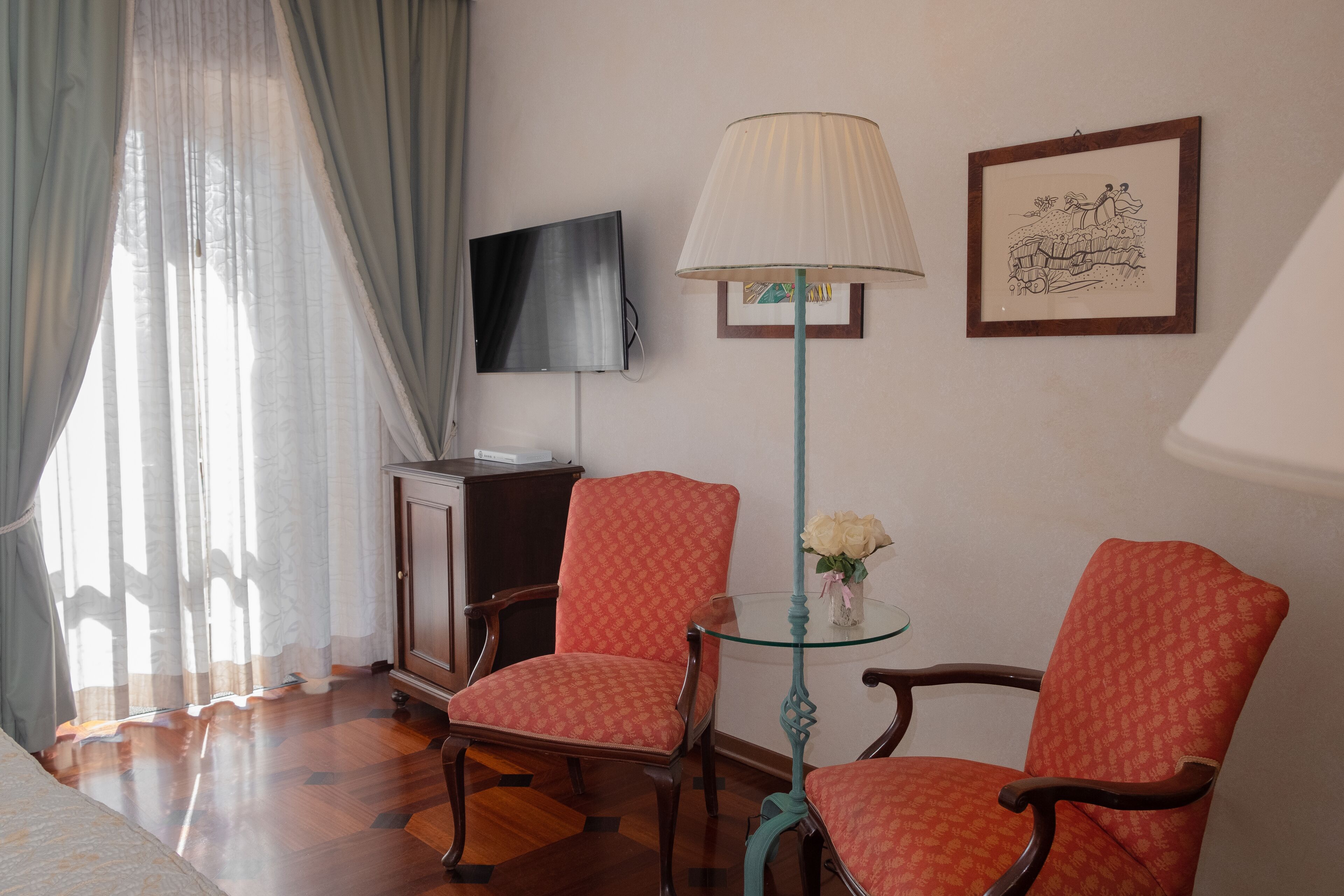 A hotel room with two red patterned chairs, a glass table with a vase of flowers, a TV on the wall, and a wooden cabinet.