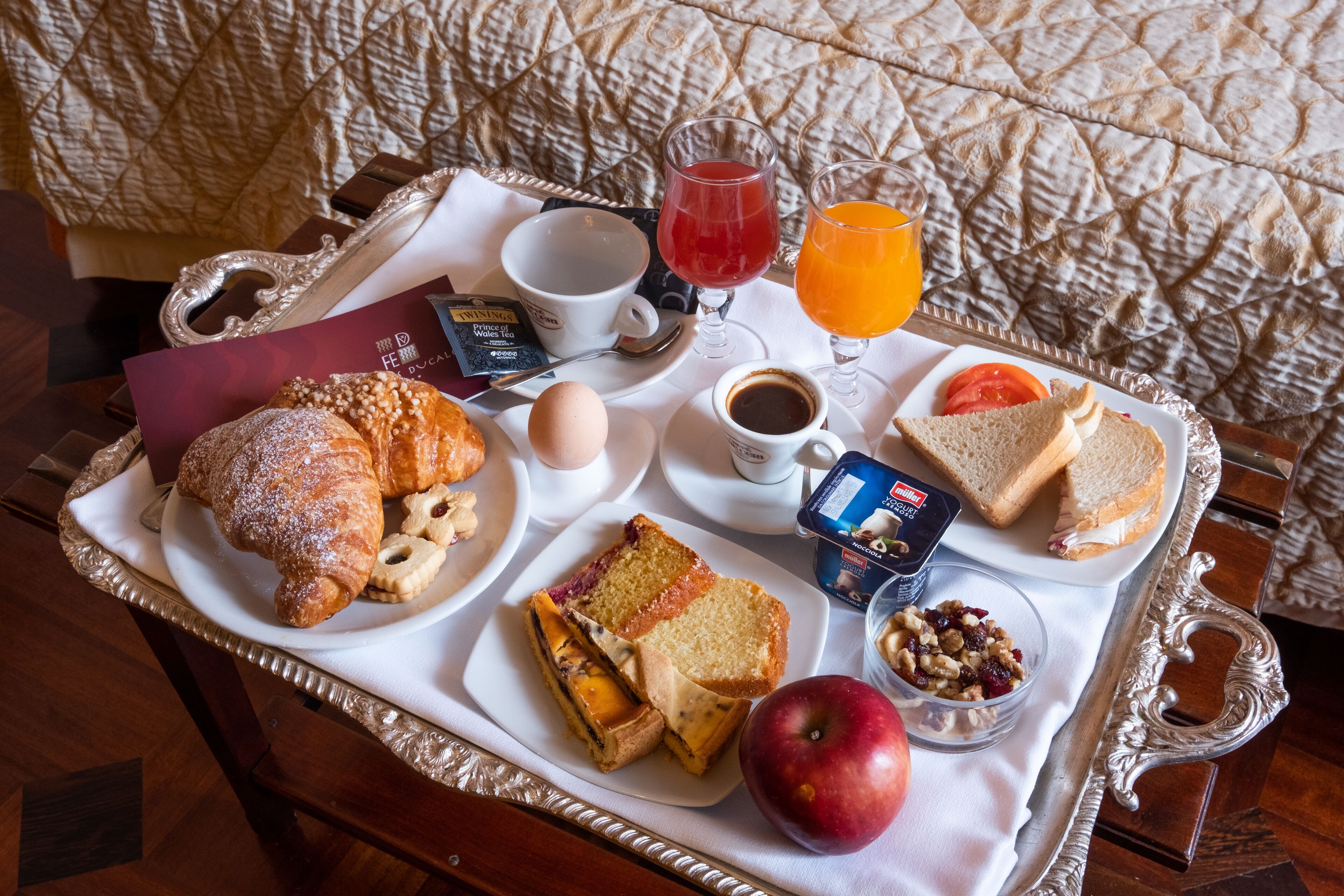 A breakfast tray with croissants, toast, an apple, a glass of orange juice, a glass of red juice, a cup of coffee, and a small container of nuts.