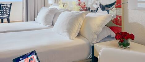 A hotel room with two beds, a bedside table with a tablet, a chair, and a vase with red flowers.