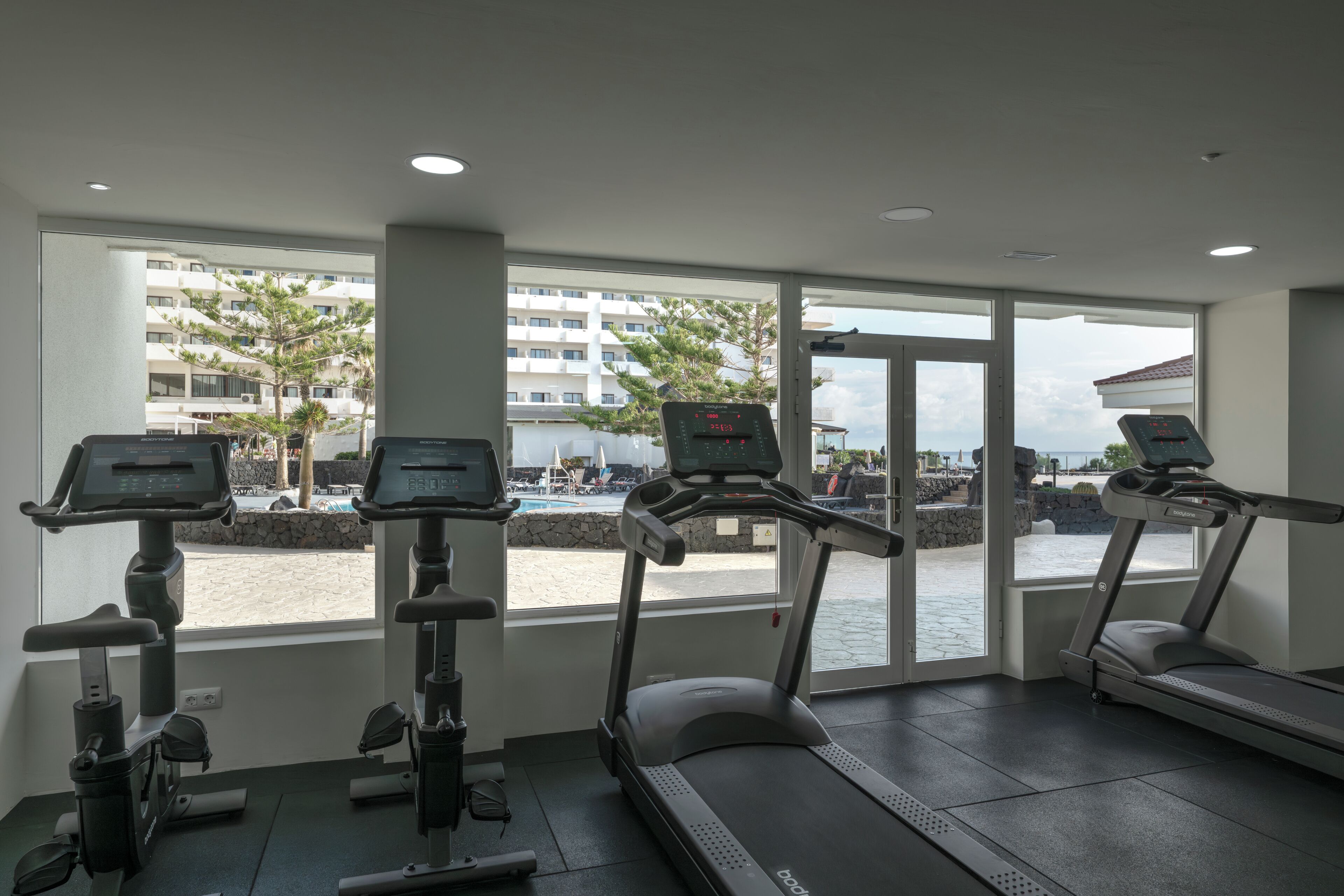 A gym with treadmills and stationary bikes, large windows with a view of a building and a body of water.