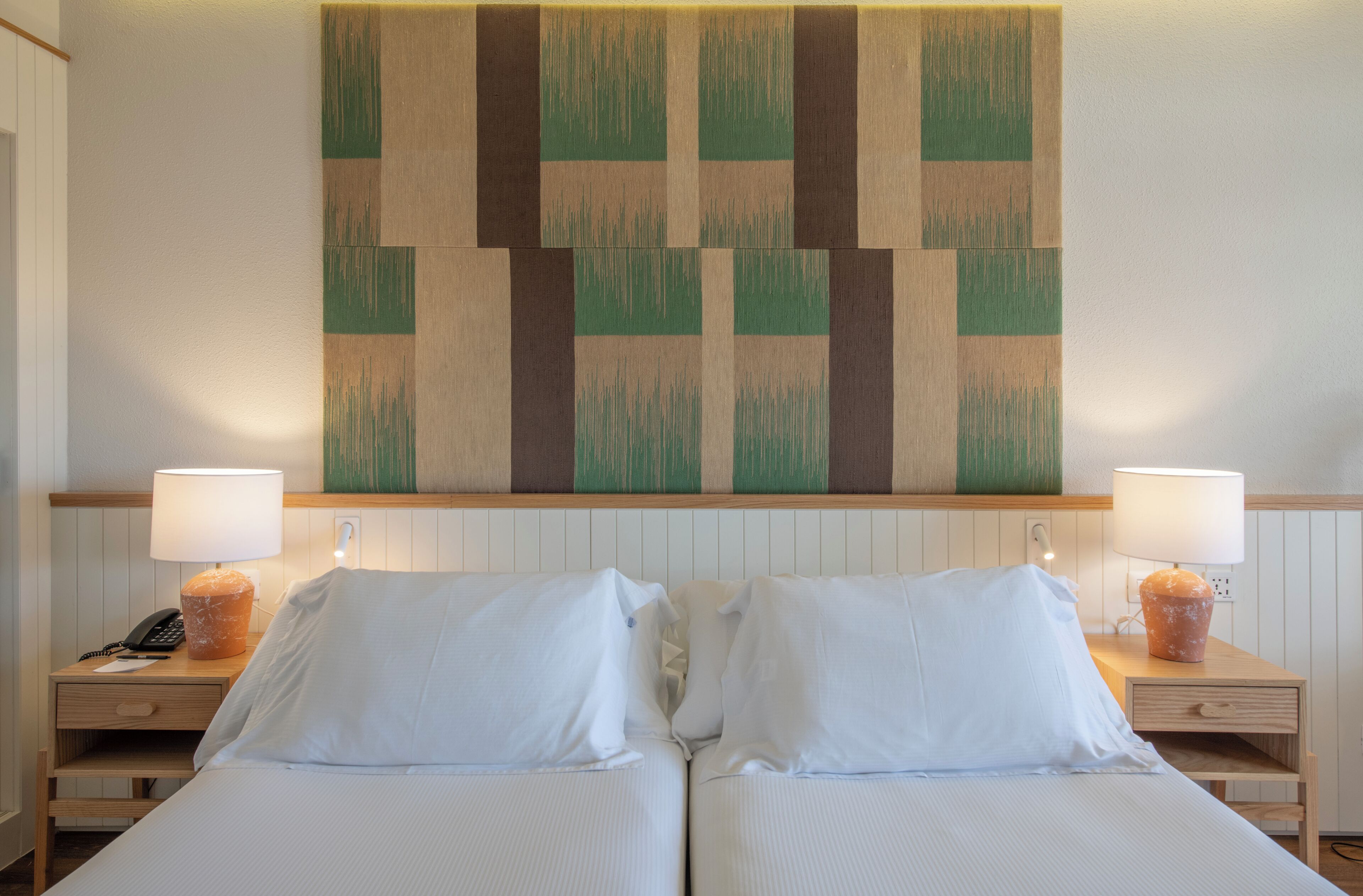 Una cama doble con sábanas blancas, dos mesitas de noche con lámparas, y una pared decorada con un diseño geométrico.