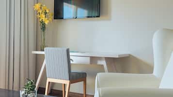 Habitación de hotel con televisión de pantalla plana montada en la pared, un escritorio blanco, una silla a rayas, un sofá blanco y una planta pequeña sobre la mesa.