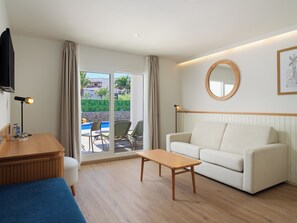 A modern hotel room with a wooden desk, a white sofa, a small wooden table, and a large window with a view of a swimming pool and greenery. - H10 White Suites (Yaiza)