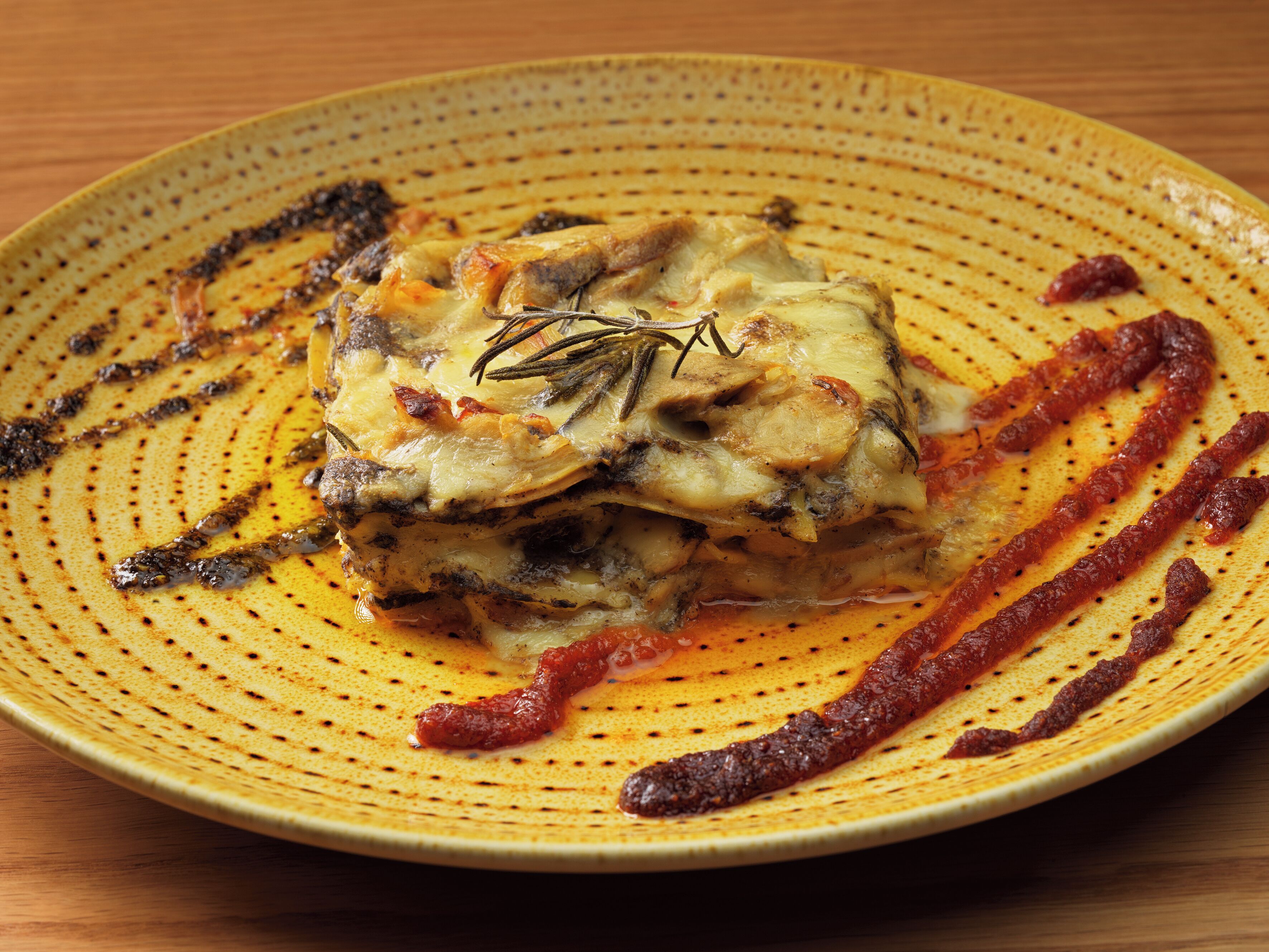 Une assiette de lasagnes nappées de fromage fondu, garnie d’une branche de romarin.