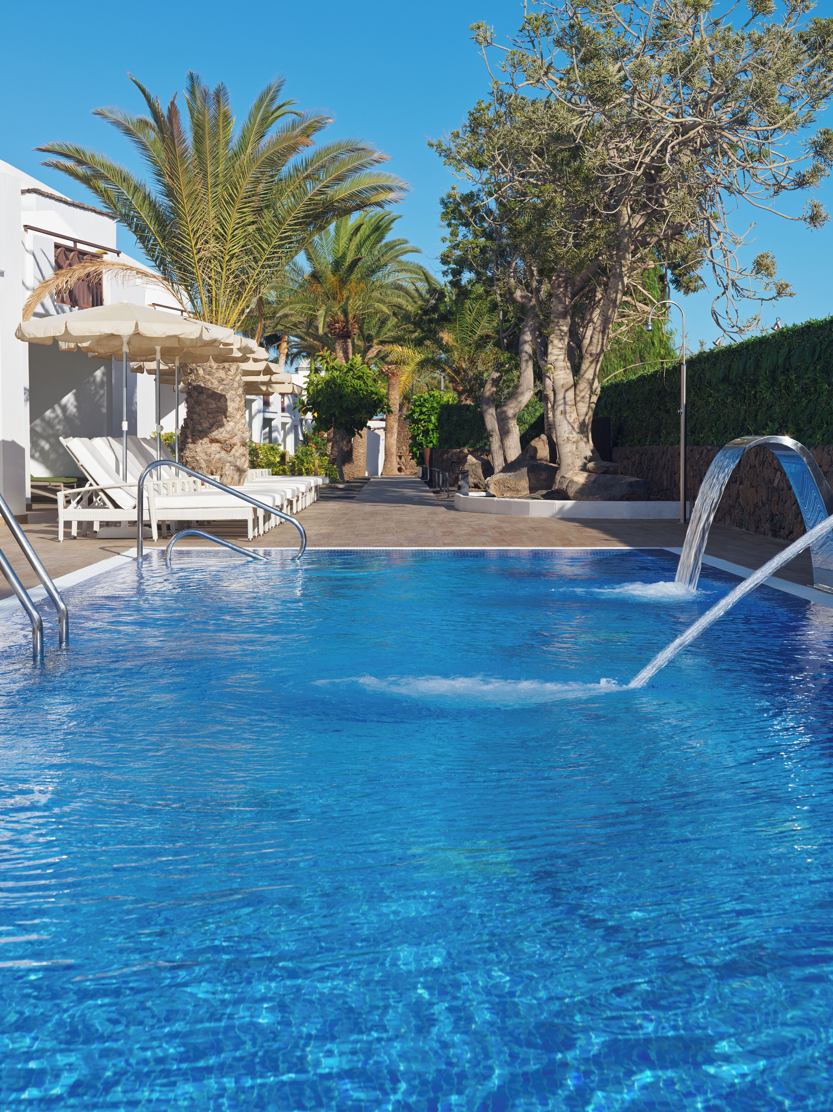 Une piscine d’un bleu éclatant, bordée de chaises longues et d’ombrelles, entourée de palmiers et d’un mur de pierre.