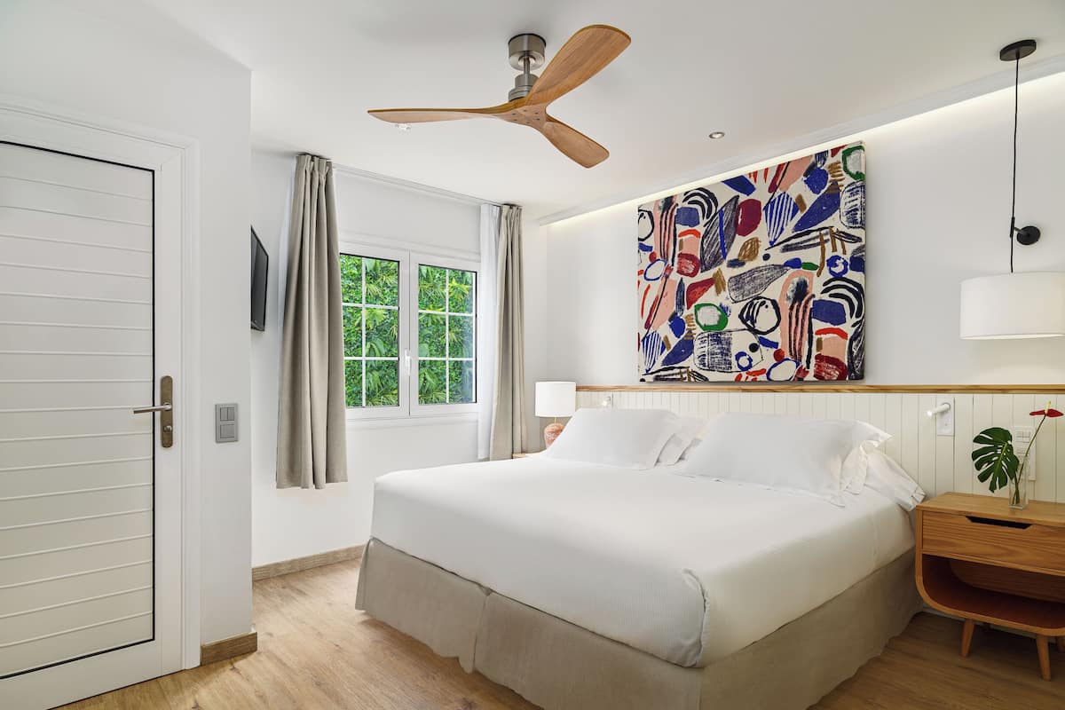 A bedroom with a bed, a ceiling fan, a window with curtains, a wall-mounted painting, and a bedside table with a lamp and a plant.