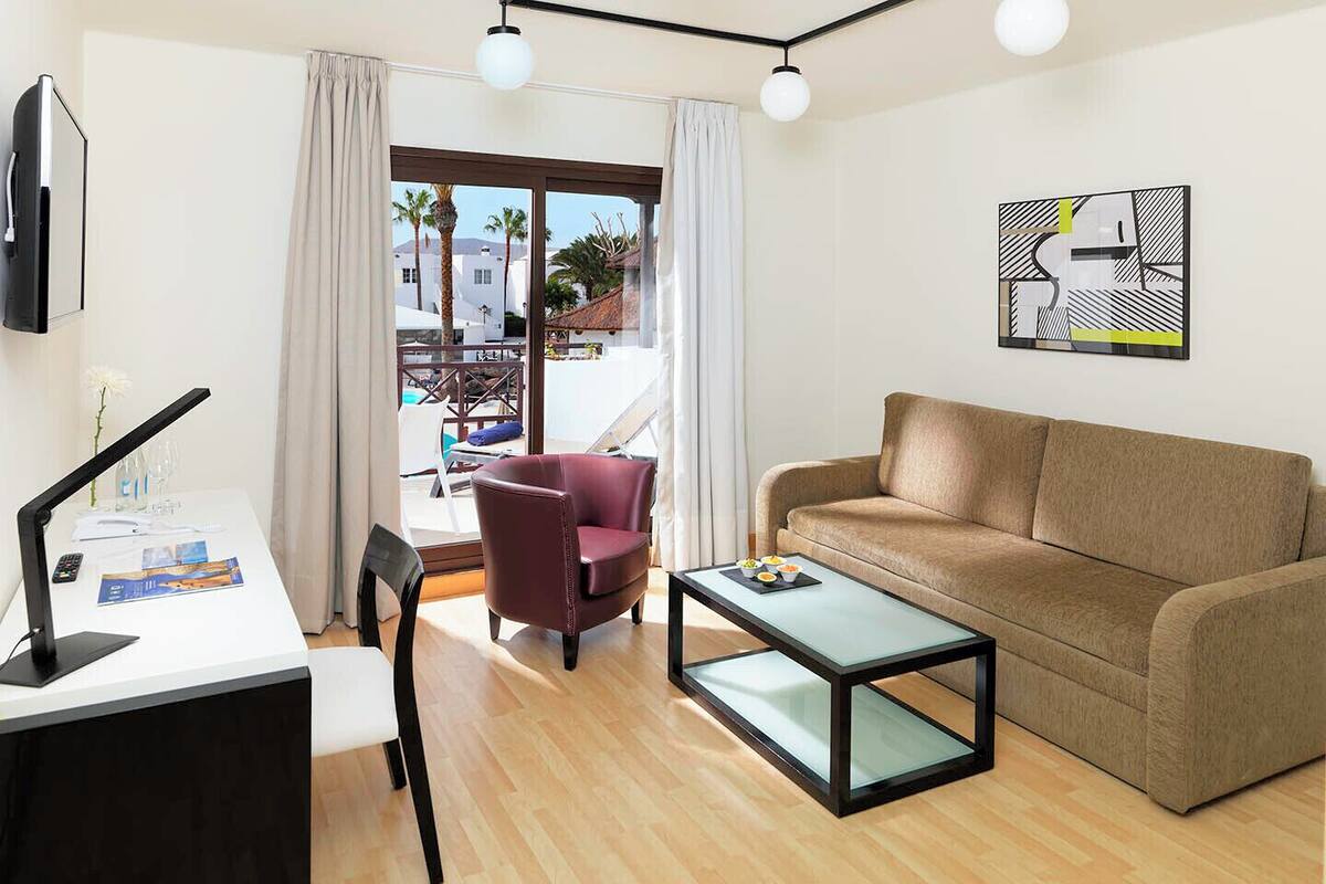 A modern living room with a sofa, armchair, and a glass coffee table. There is a desk with a lamp and a remote control. A painting is on the wall, and a window with curtains is visible.
