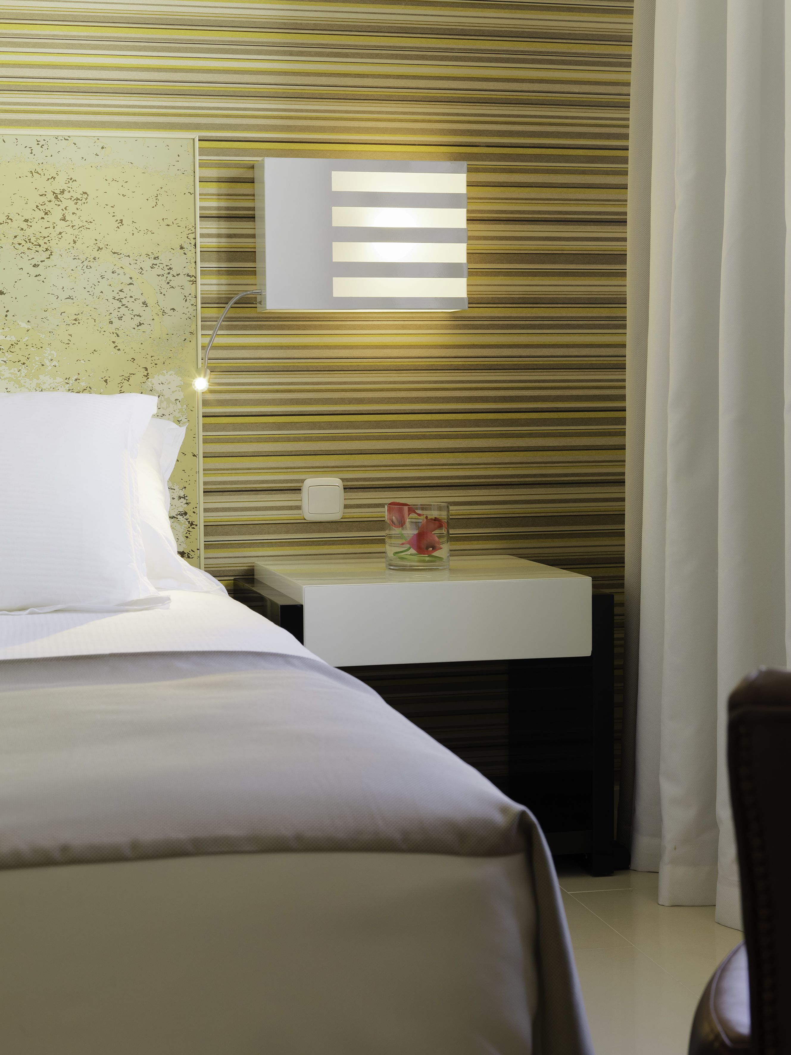 Una habitación de hotel con una cama, una mesita de noche con un vaso rojo, un espejo montado en la pared y un diseño de rayas en la pared.