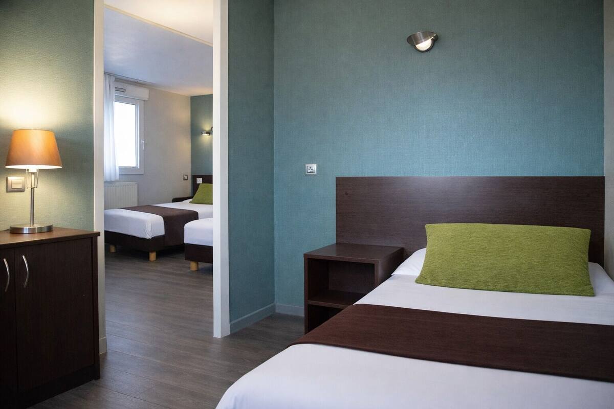 Ein Hotelzimmer mit zwei Betten, einem Nachttisch, einer Lampe und Blick auf ein anderes Zimmer.