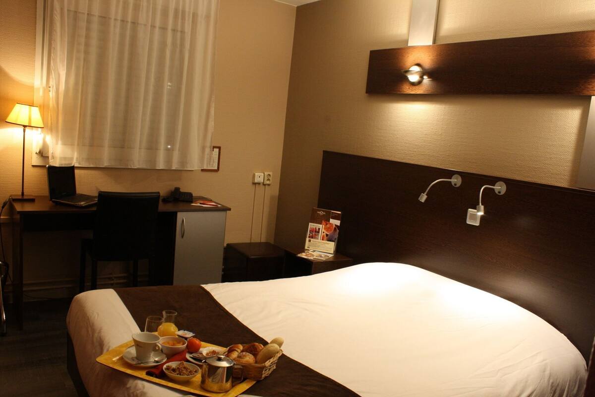 Ein Hotelzimmer mit Bett, Schreibtisch mit Laptop, kleinem Kühlschrank, Nachttisch mit Lampe und Frühstückswagen.