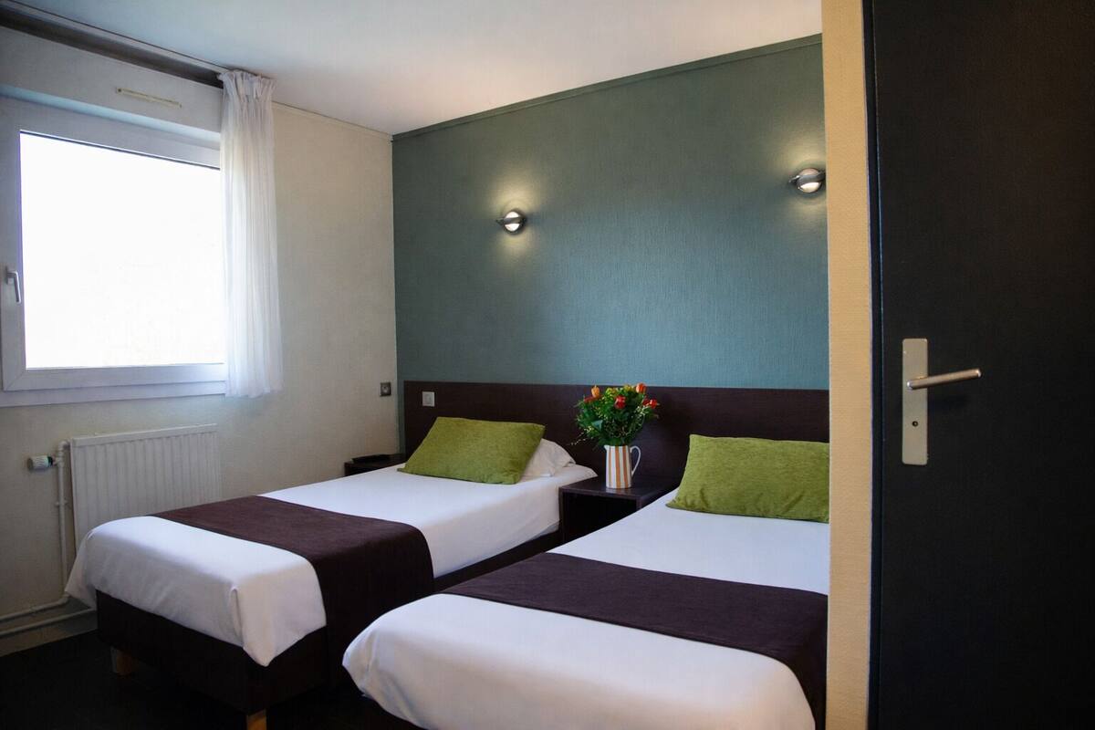 Ein Hotelzimmer mit zwei Betten, einem Fenster mit Vorhängen, einem Heizkörper und einem Nachttisch mit Blumen.