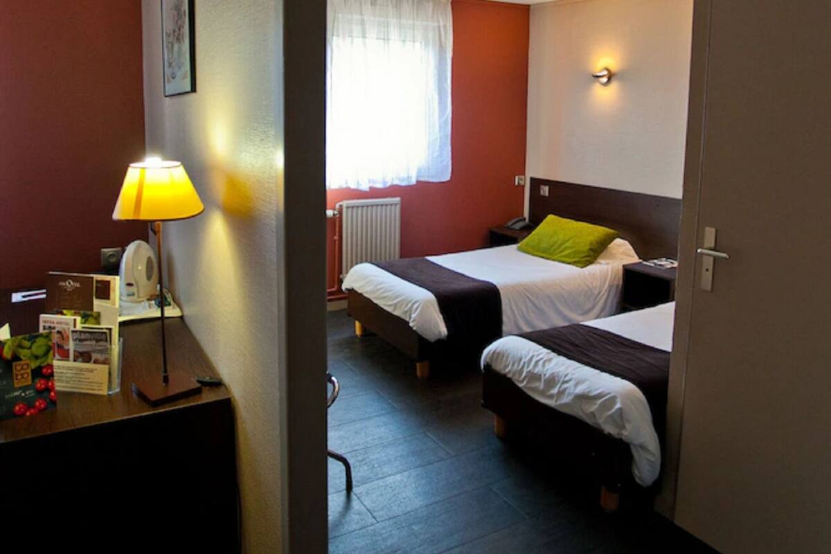 Ein Hotelzimmer mit zwei Betten, einem Schreibtisch mit Lampe, einem Fenster mit Vorhängen und einer Wandleuchte.