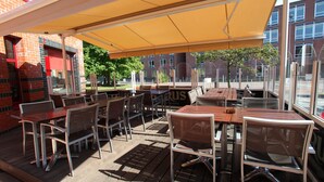 Une terrasse extérieure avec des tables et des chaises en bois, un auvent jaune et un bâtiment en briques en arrière-plan.