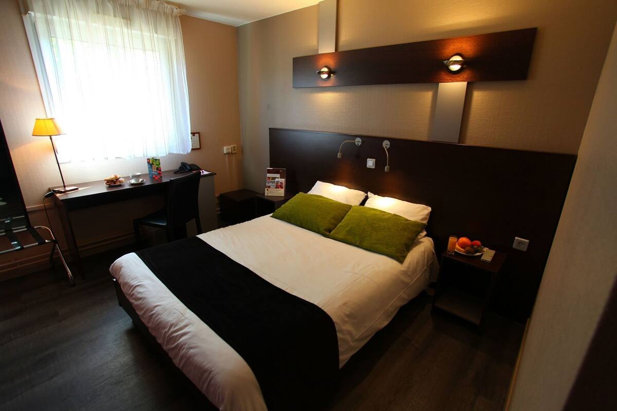 Ein Hotelzimmer mit Bett, Schreibtisch, Stuhl, Nachttisch, Lampe und einem Fenster mit Vorhängen.