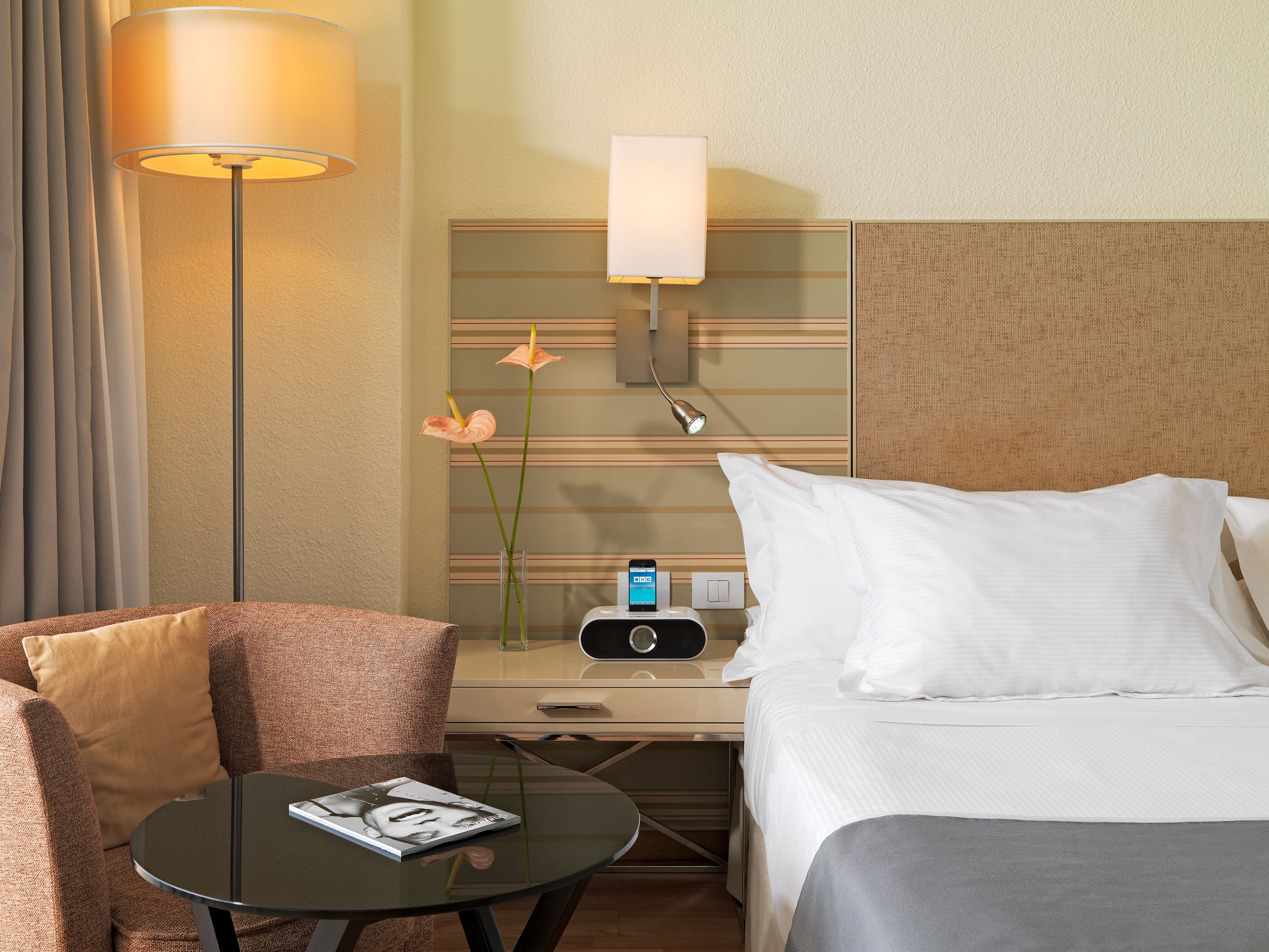 Una habitación de hotel con cama, mesita de noche, lámpara, un jarrón con flores, una mesa con revista y un radio incorporado a la pared.